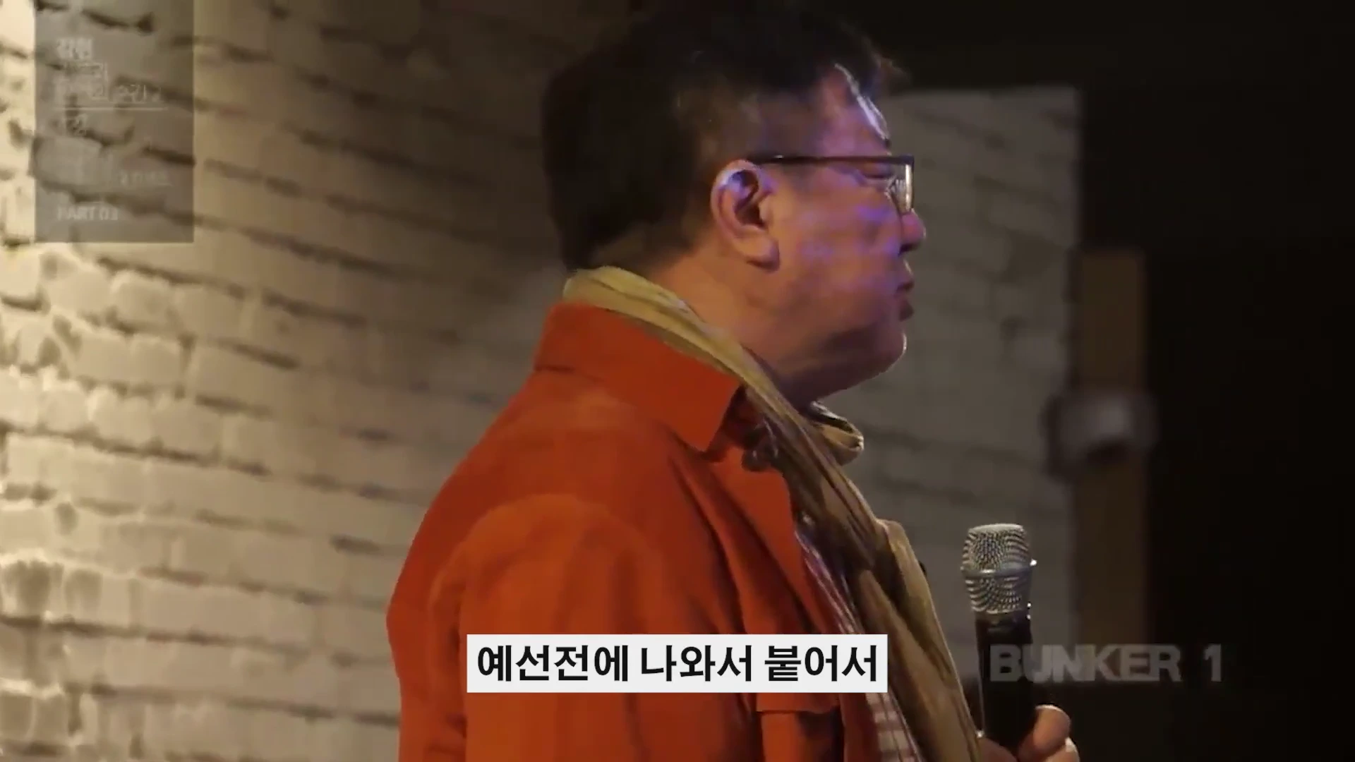 안경을 쓴 남성이 마이크를 들고 벽돌 벽을 배경으로 강연하고 있는 모습