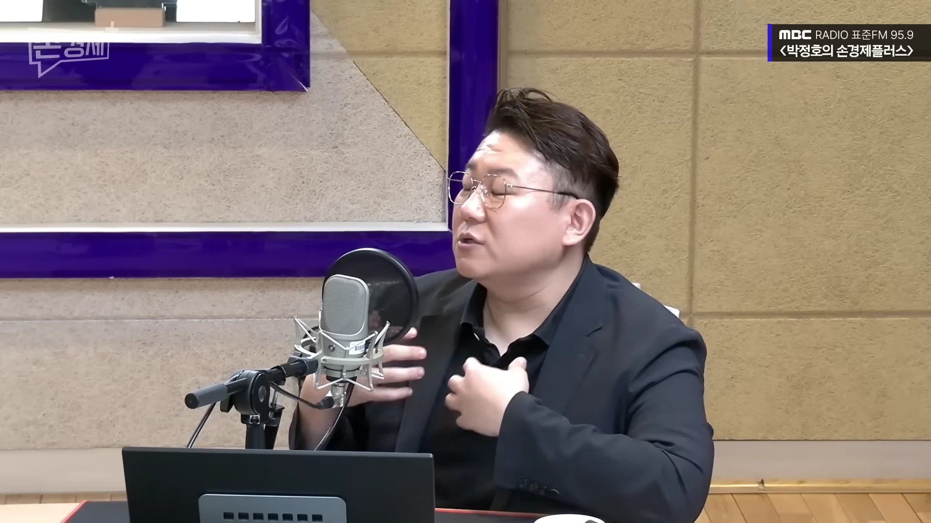 안경을 쓴 남성이 라디오 스튜디오에서 마이크 앞에 앉아 손짓하며 이야기하고 있다.