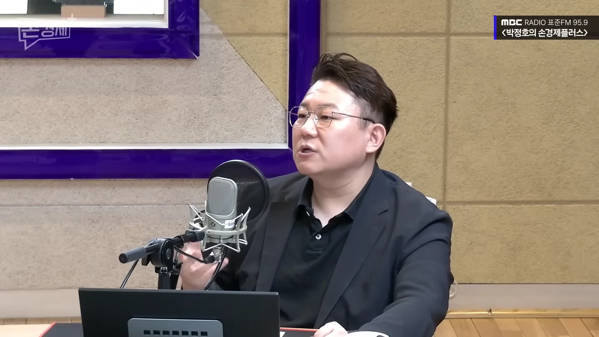 안경을 쓴 남성이 라디오 스튜디오에서 마이크 앞에 앉아 이야기를 나누고 있다.