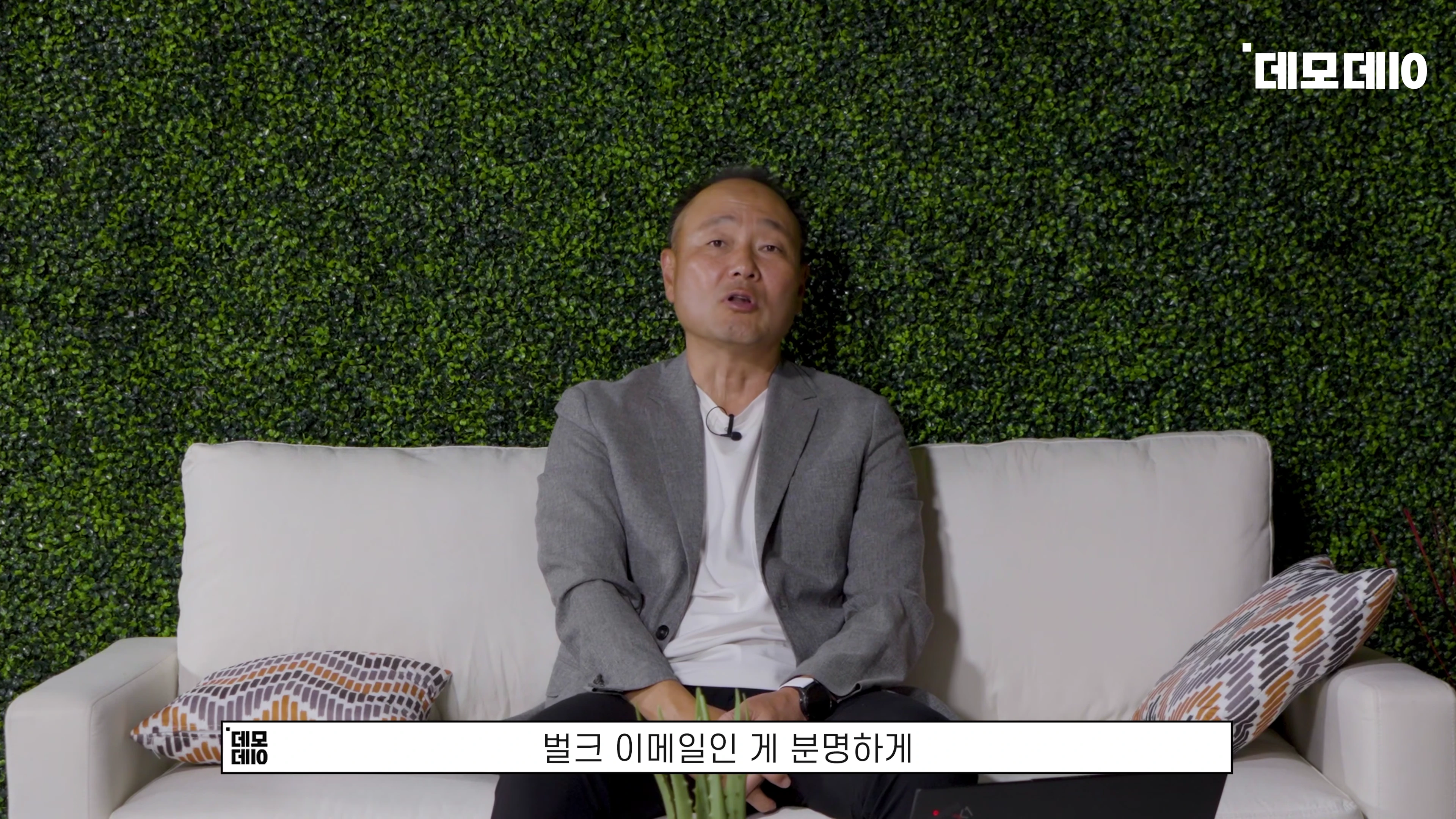 초록색 인조 잔디 벽을 배경으로 흰색 소파에 앉아 이야기하는 중년 남성