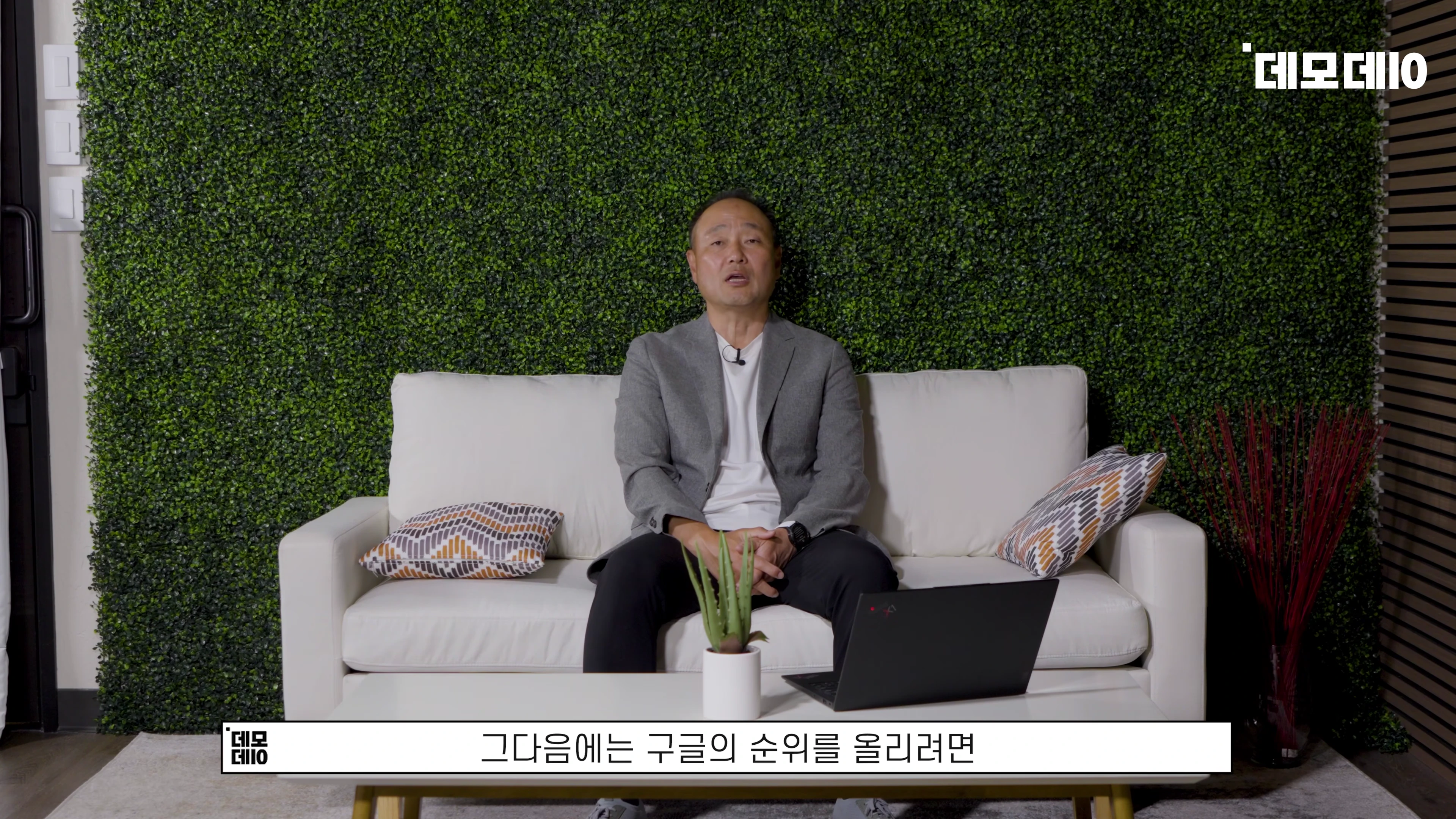 초록색 인조 식물 벽을 배경으로 흰색 소파에 앉아 이야기하는 중년 남성