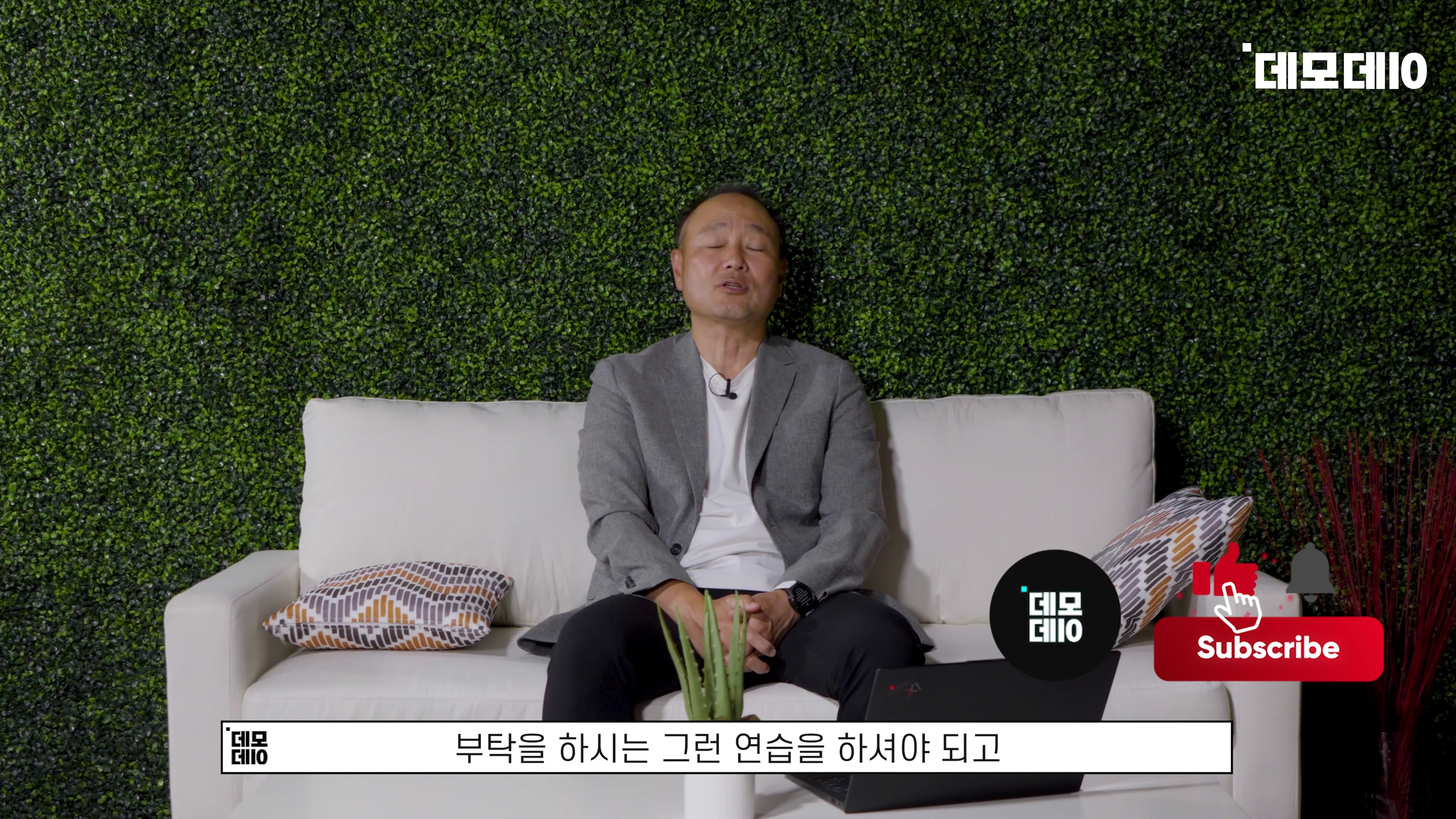 초록색 인조 잔디 벽을 배경으로 흰색 소파에 앉아 이야기하는 중년 남성