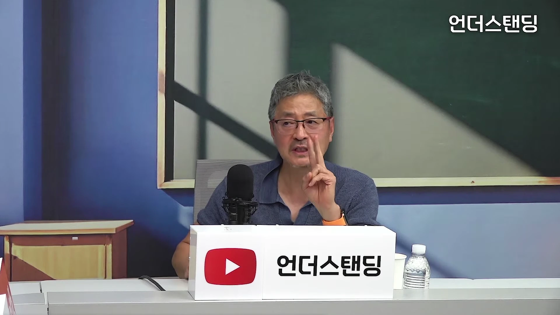 안경을 쓴 중년 남성이 스튜디오 책상에 앉아 손가락으로 숫자 2를 표시하며 이야기하고 있다.