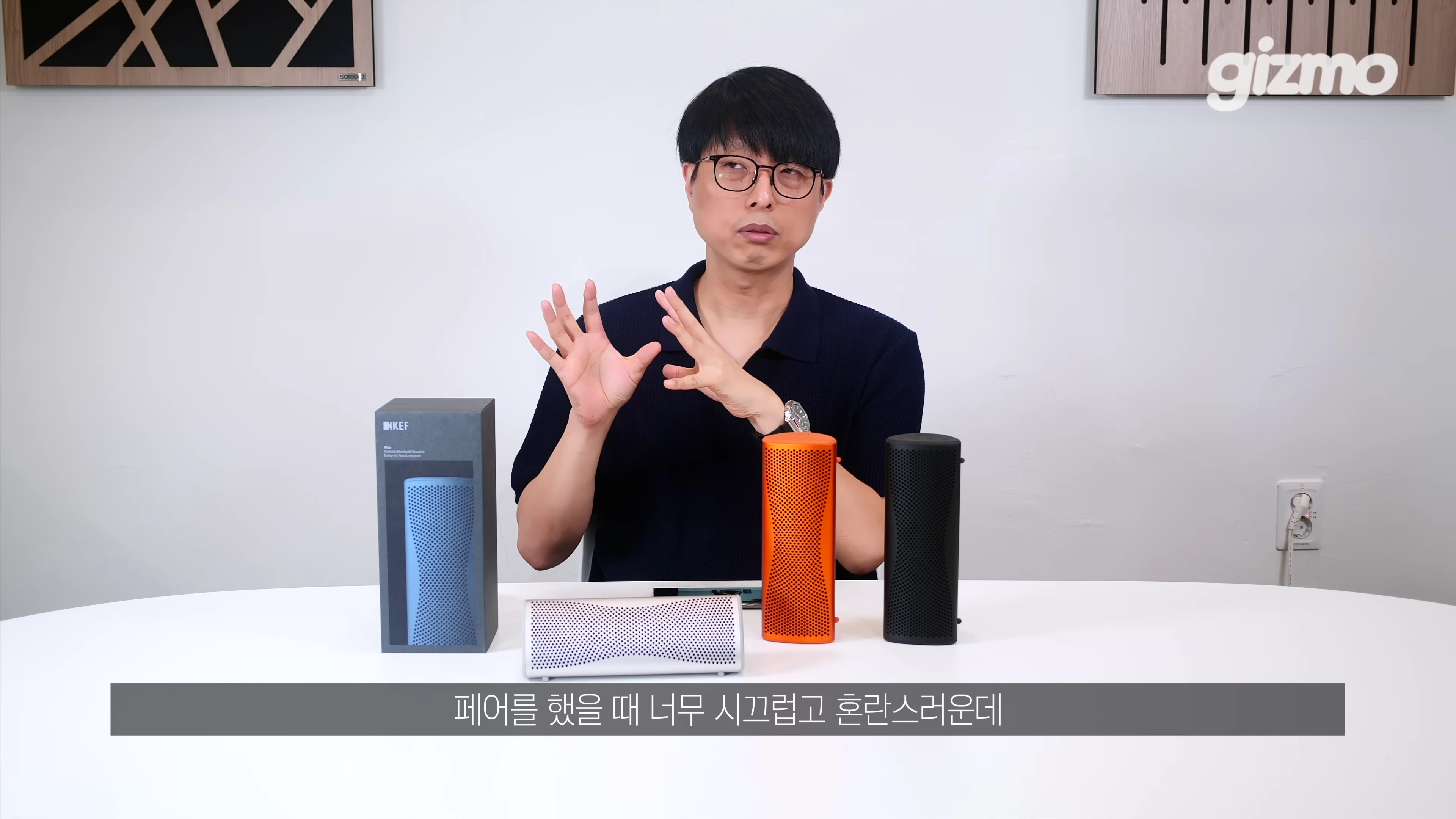 스튜디오에서 안경을 쓴 남성이 손짓하며 KEF 휴대용 스피커 제품들을 설명하고 있다.