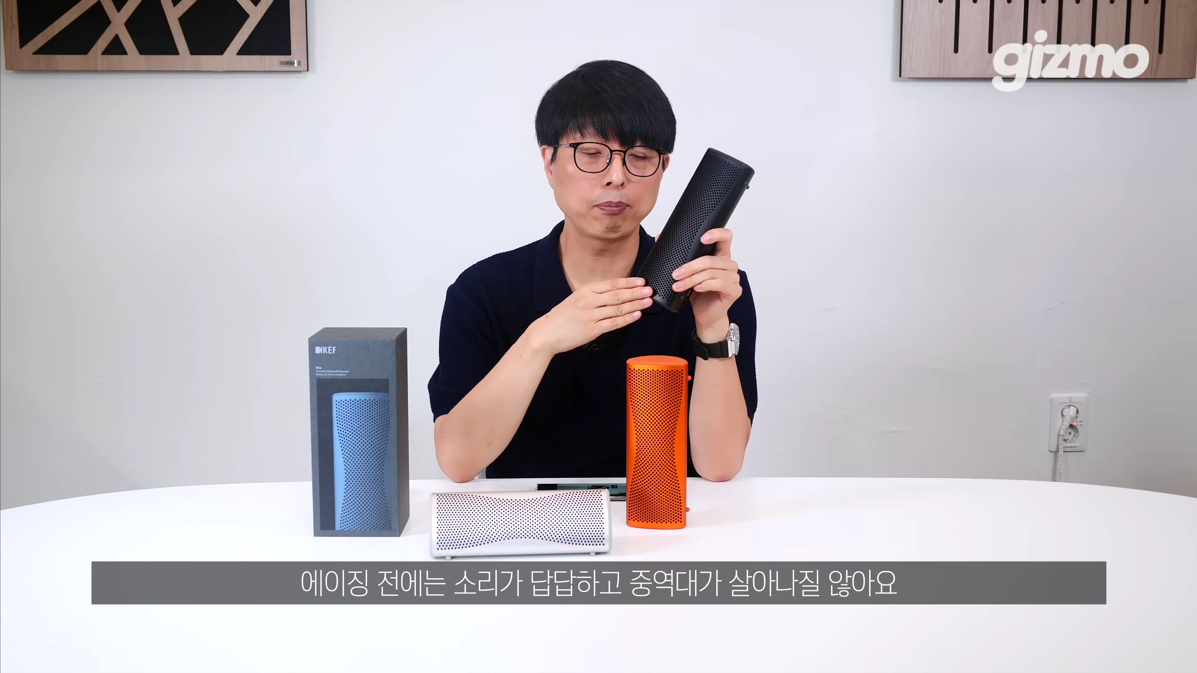 안경을 쓴 남성이 테이블 위에 놓인 휴대용 스피커들을 보여주며 설명하고 있다.
