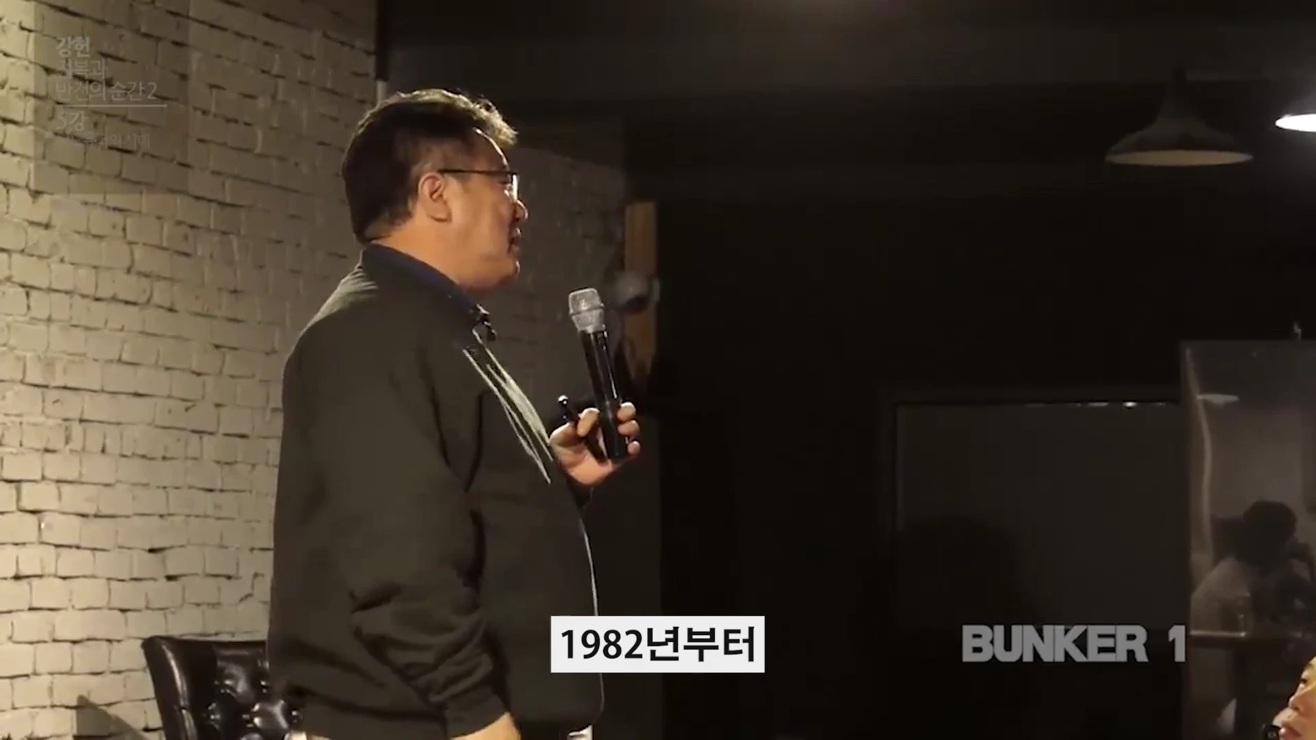 어두운 실내에서 마이크를 들고 강연하는 중년 남성의 옆모습