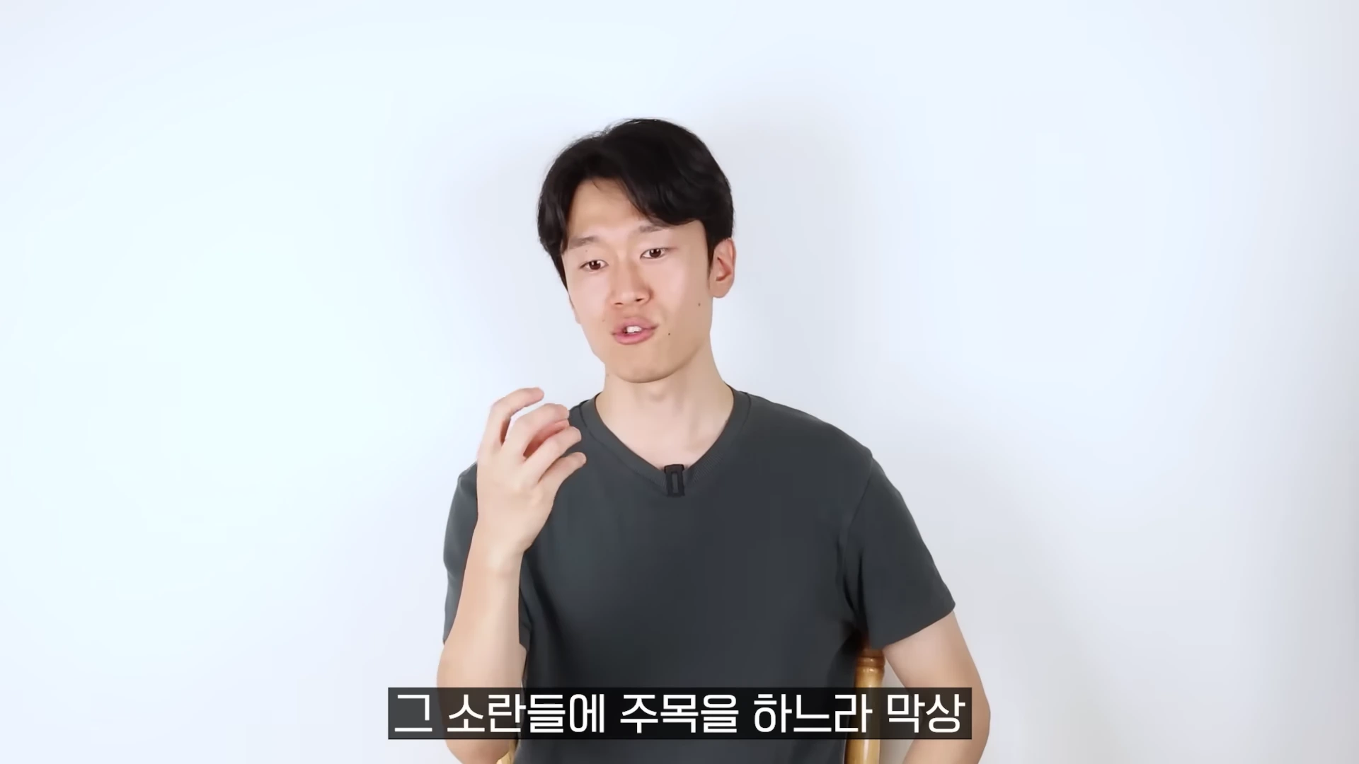 흰 배경 앞에서 어두운 회색 티셔츠를 입은 남성이 손짓하며 이야기하는 모습