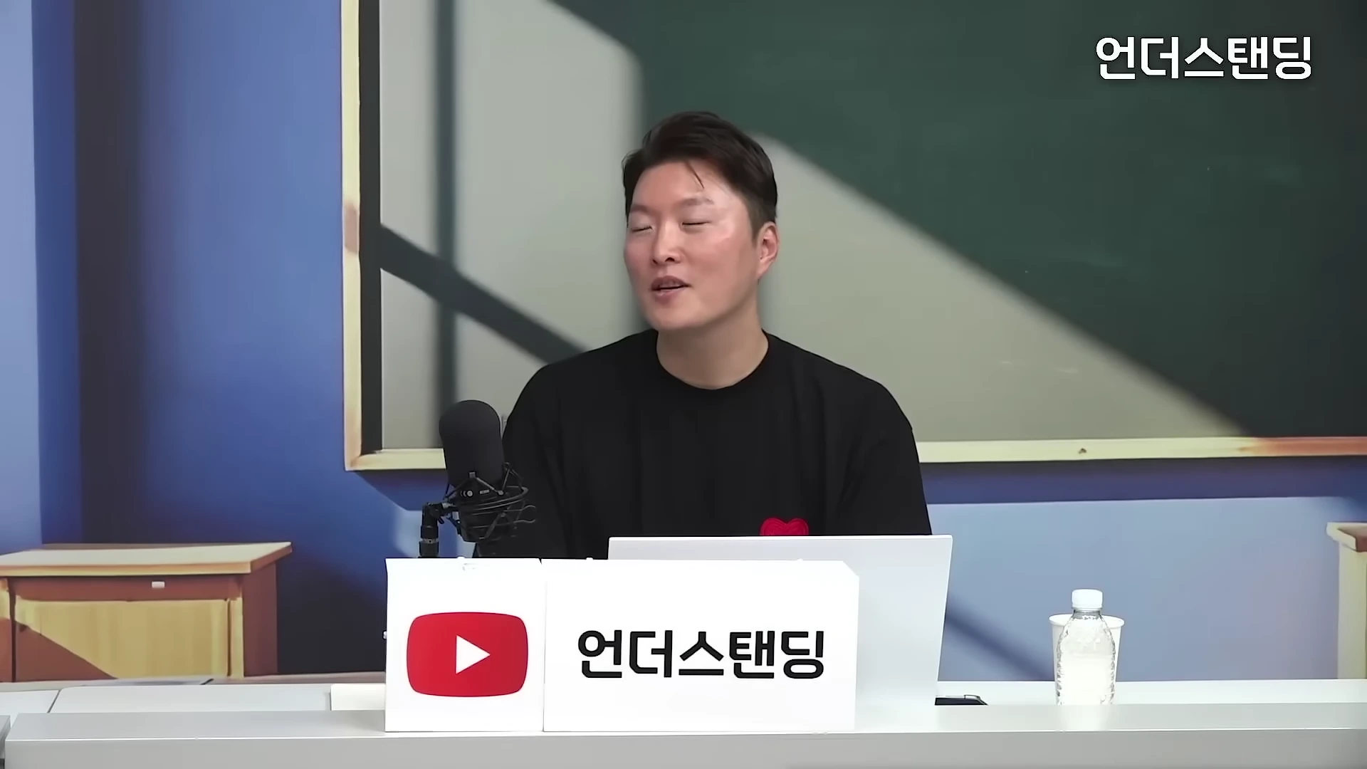 검은색 티셔츠를 입은 남성이 스튜디오 책상 앞에 앉아 마이크를 앞에 두고 이야기를 나누고 있다.