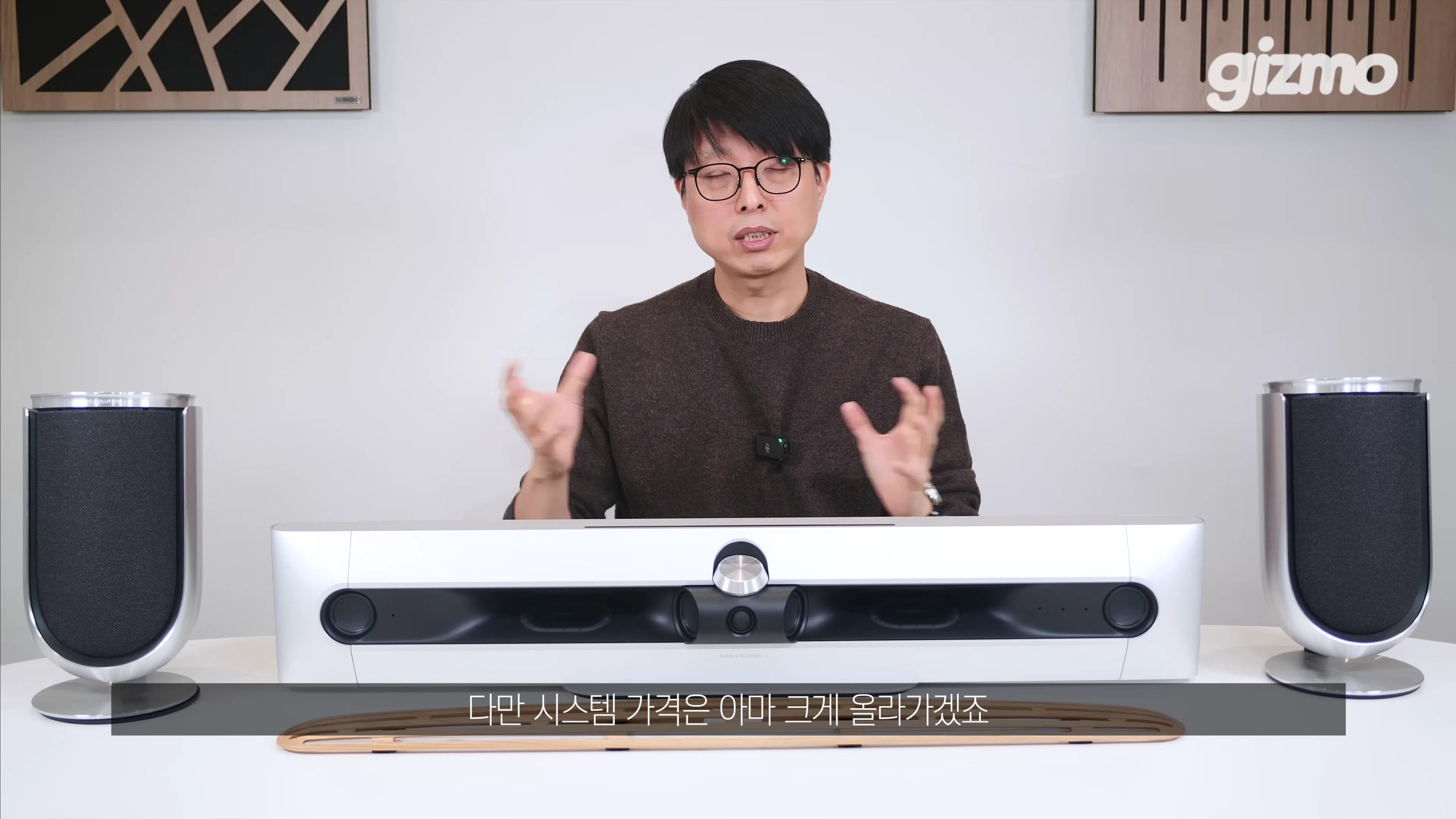 안경을 쓴 남성이 테이블 위에 놓인 사운드바와 스피커를 앞에 두고 손짓하며 설명하고 있다.