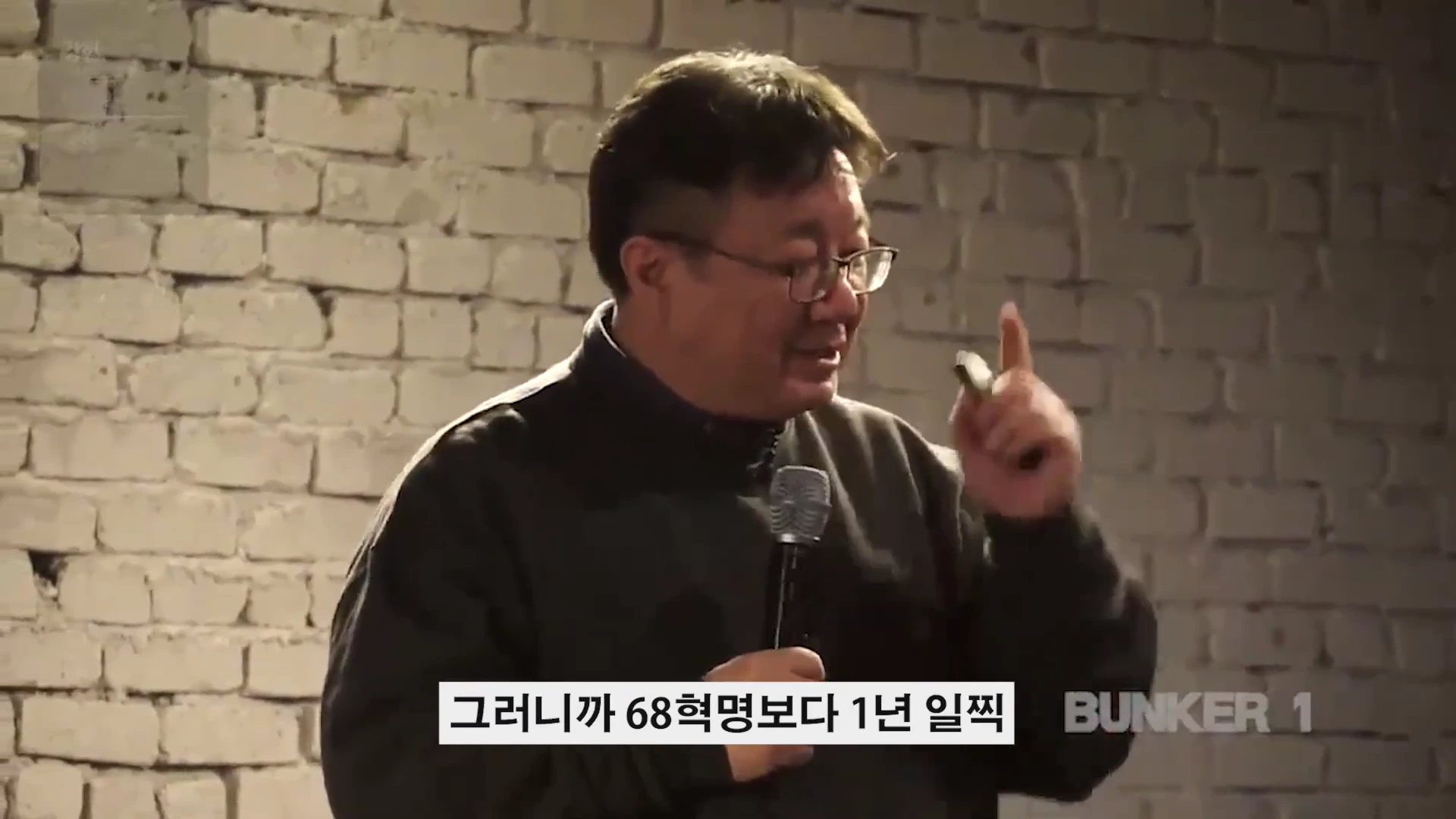 안경을 쓴 중년 남성이 마이크를 들고 손가락을 들어 올리며 강연하는 모습