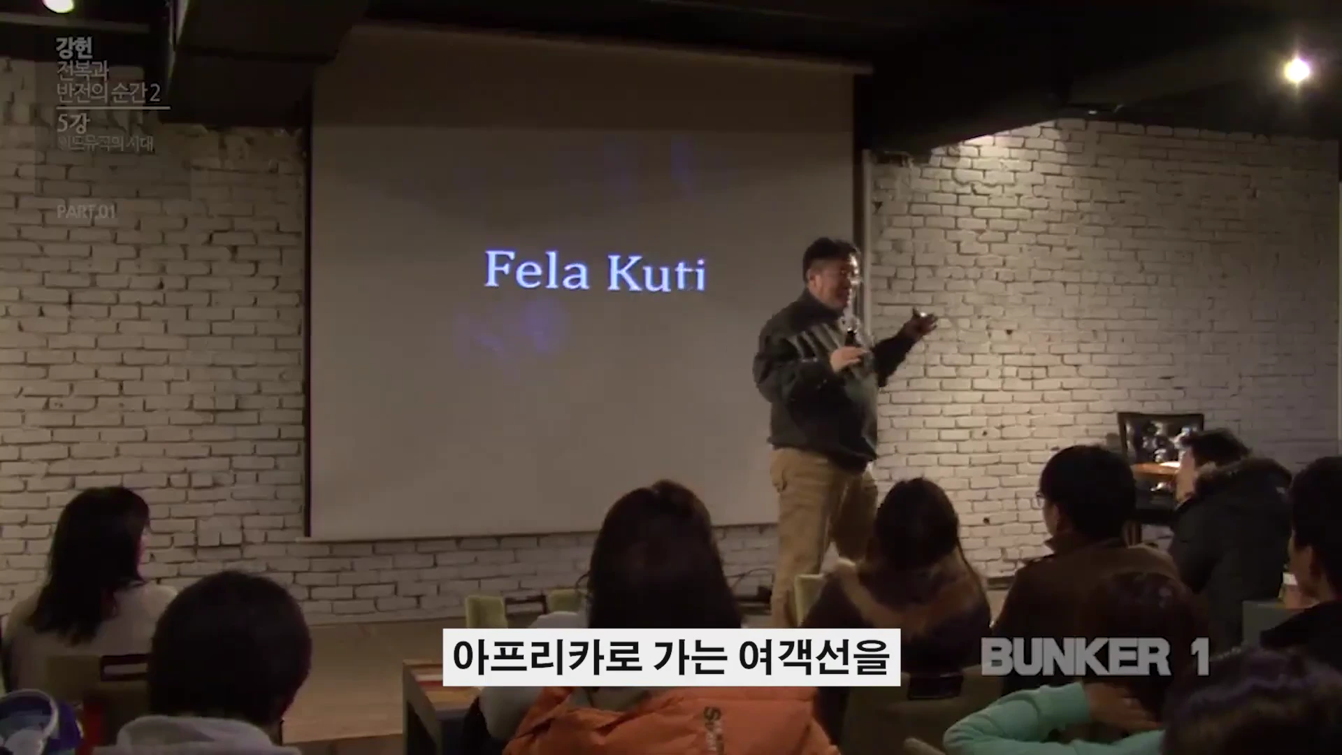 강연자가 스크린에 'Fela Kuti'라는 이름이 띄워진 무대에서 청중을 향해 이야기하고 있다.
