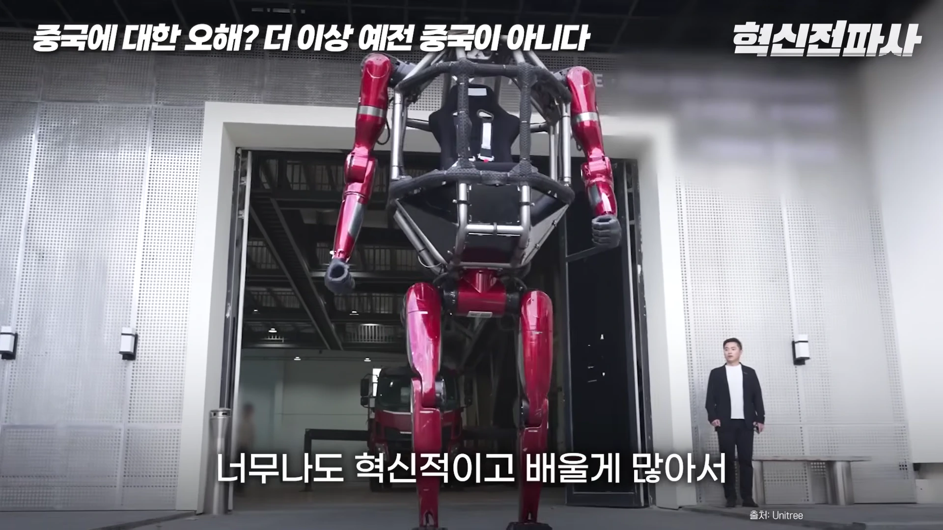 실내에서 사람이 탑승할 수 있는 붉은색의 거대한 휴머노이드 로봇이 서 있고, 옆에 한 남성이 서 있는 영상 화면