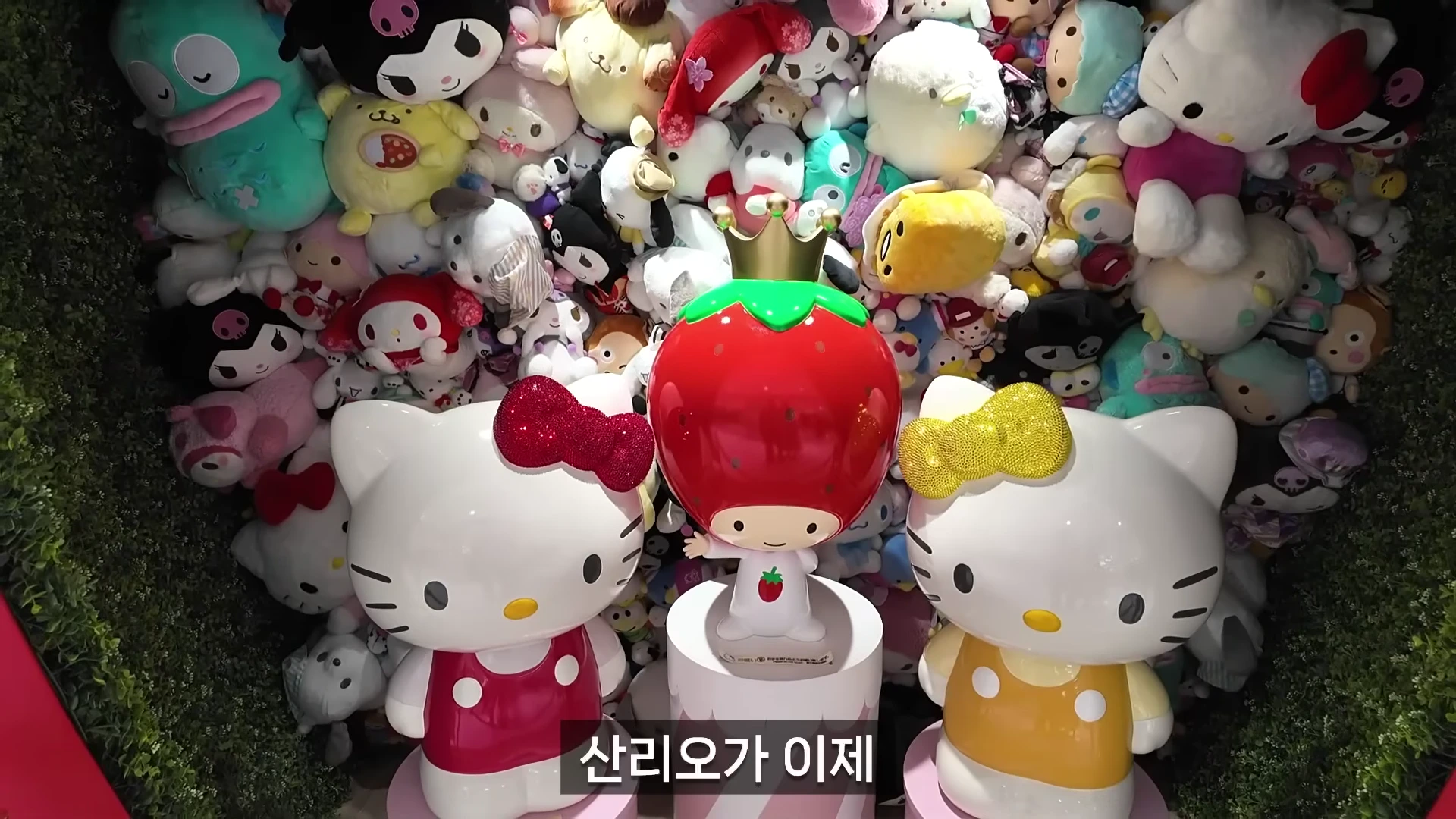 다양한 산리오 캐릭터 인형들이 벽면을 가득 채우고 있고, 앞쪽에는 헬로키티 조형물 두 개와 딸기 모자를 쓴 캐릭터 조형물이 놓여 있습니다.