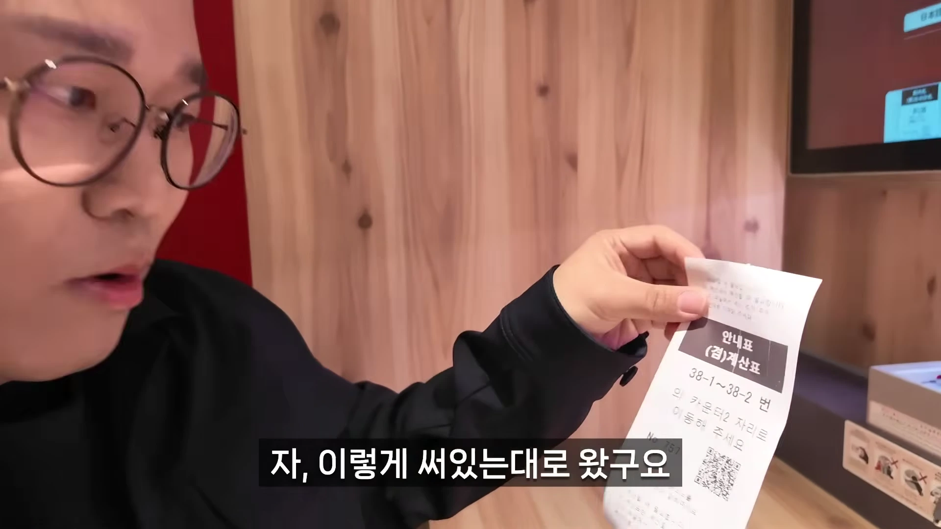 안경을 쓴 남성이 식당에서 테이블 번호가 적힌 안내표를 손에 들고 보여주고 있다.