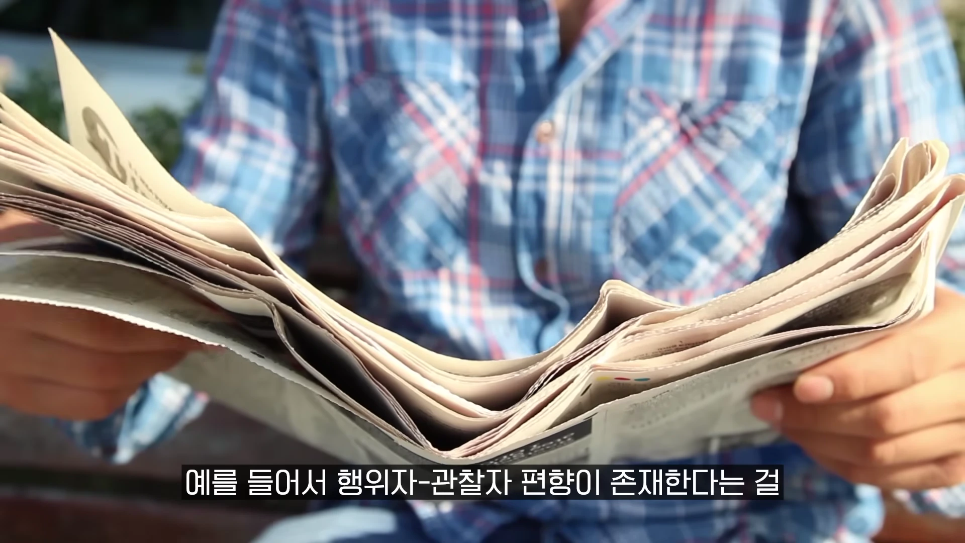 파란색 체크무늬 셔츠를 입은 사람이 신문을 펼쳐 들고 있는 모습