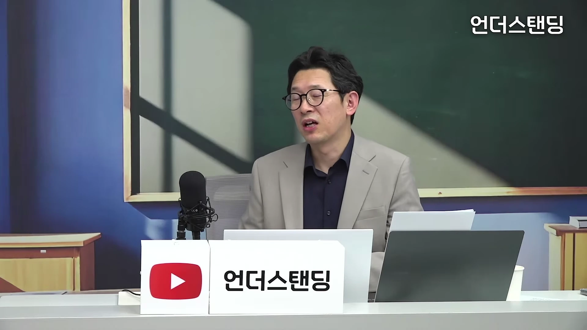 안경을 쓴 남성이 스튜디오 책상에 앉아 마이크를 앞에 두고 이야기를 나누고 있다.