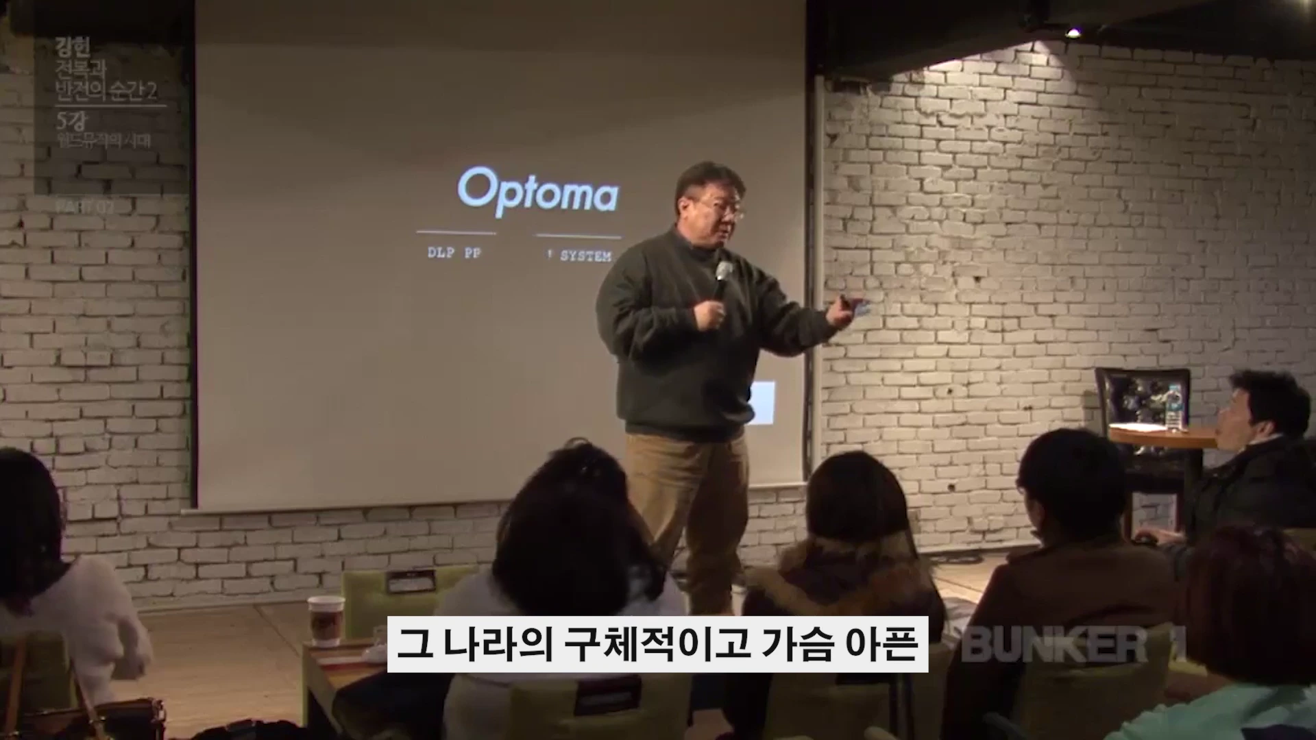 강연자가 흰 벽돌 배경의 무대에서 마이크를 들고 청중을 향해 이야기하고 있다.