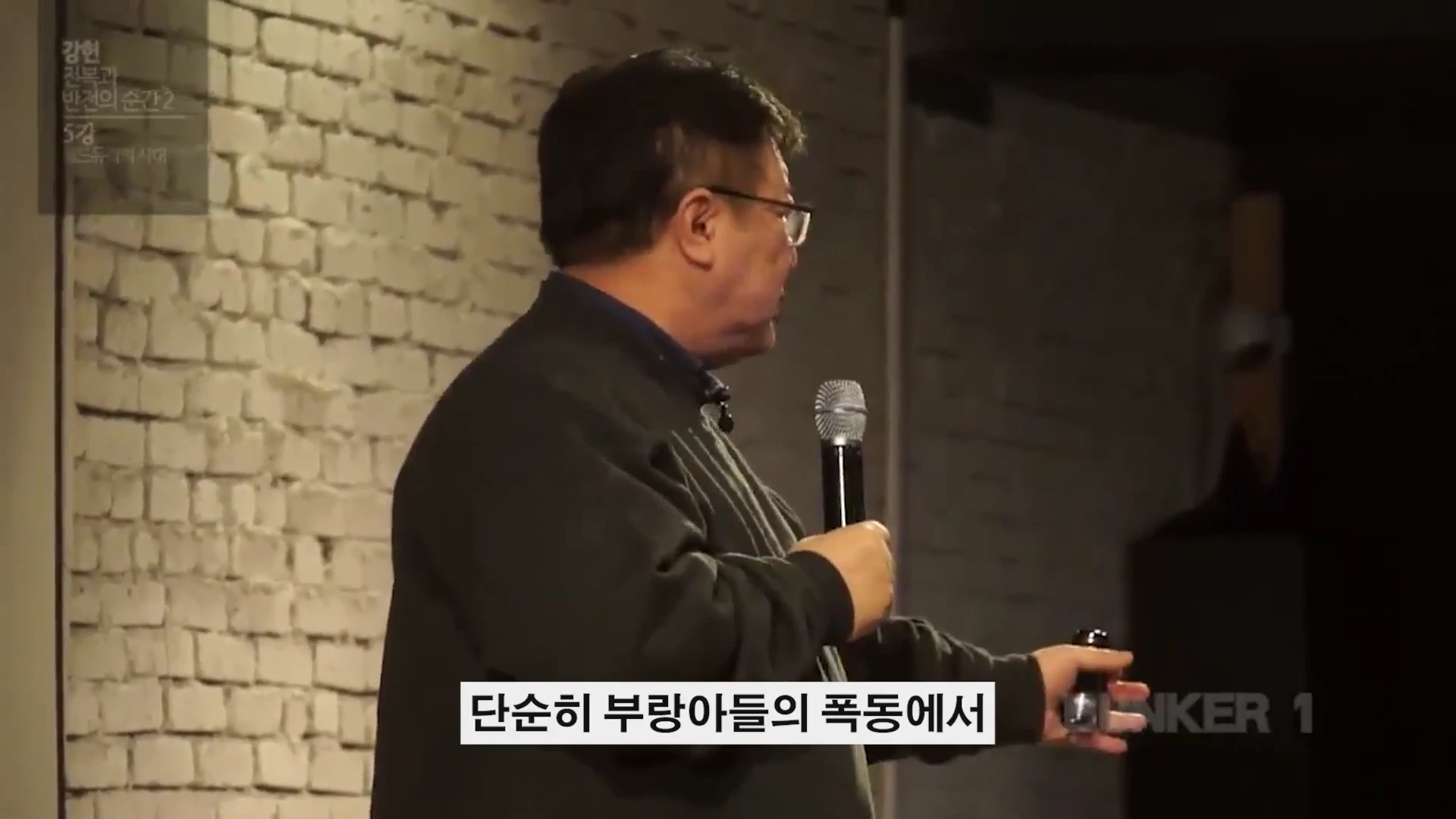 안경을 쓴 중년 남성이 마이크를 들고 흰 벽돌 벽을 배경으로 강연을 하고 있다.