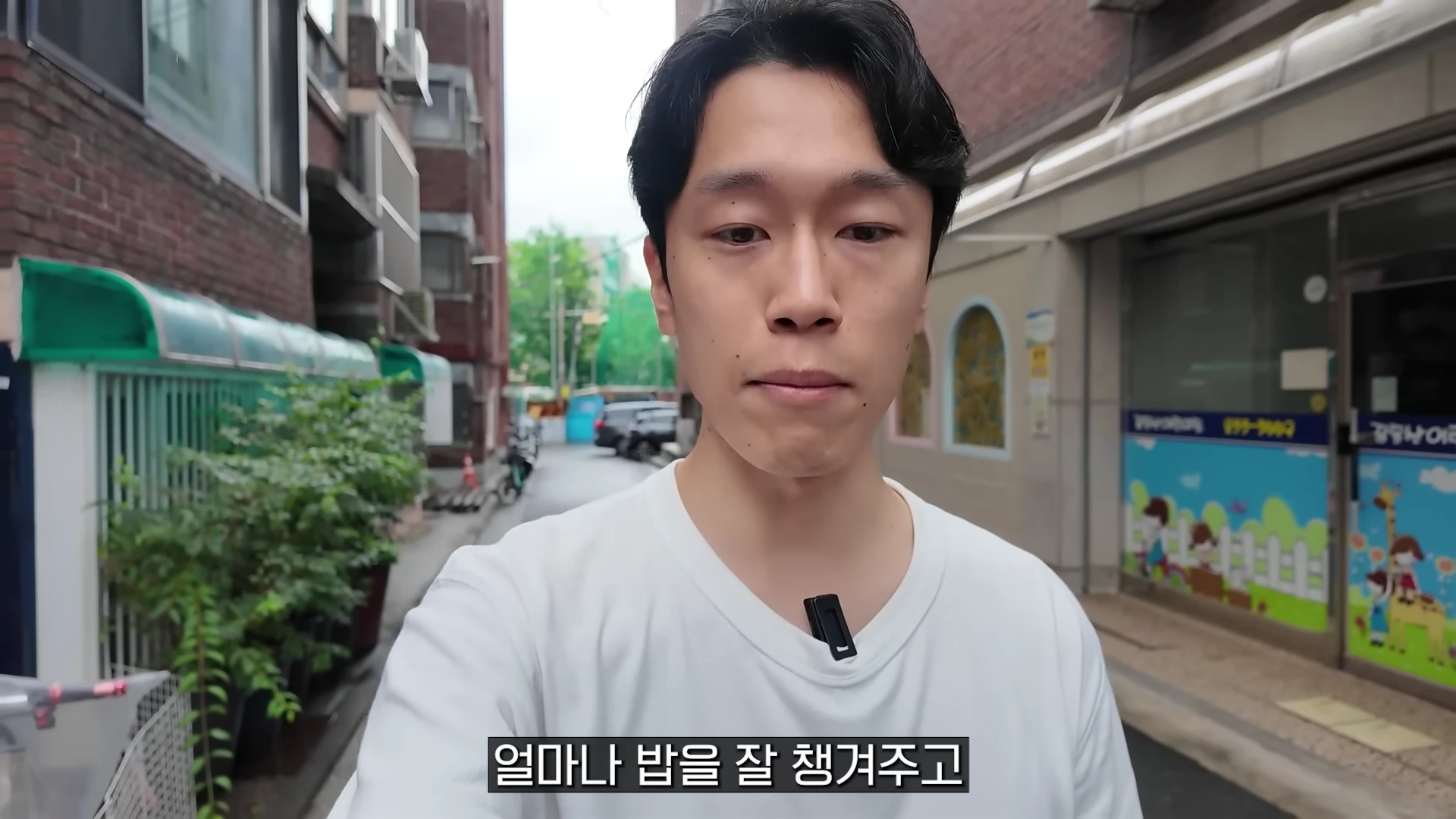 흰색 티셔츠를 입은 남성이 골목길에서 카메라를 응시하며 이야기하고 있다.