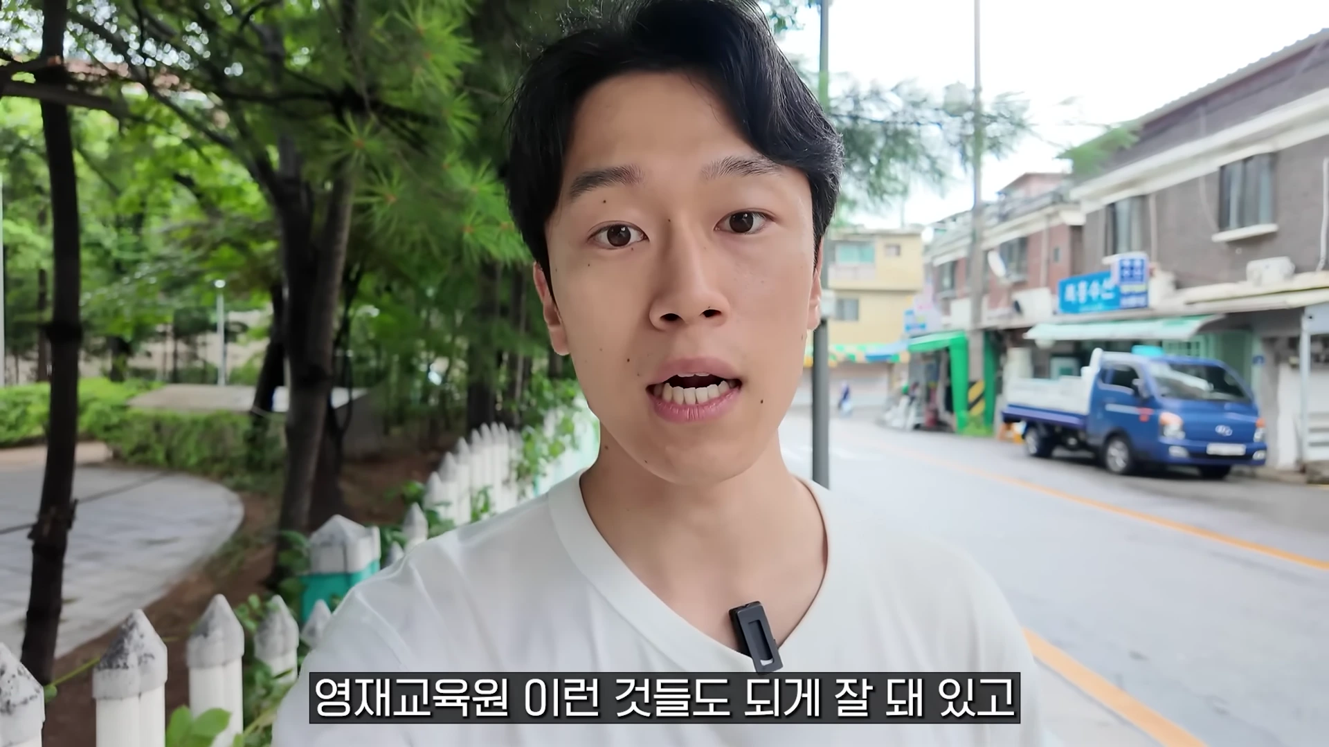 흰색 티셔츠를 입은 남성이 야외 거리에서 카메라를 보며 이야기하고 있다.