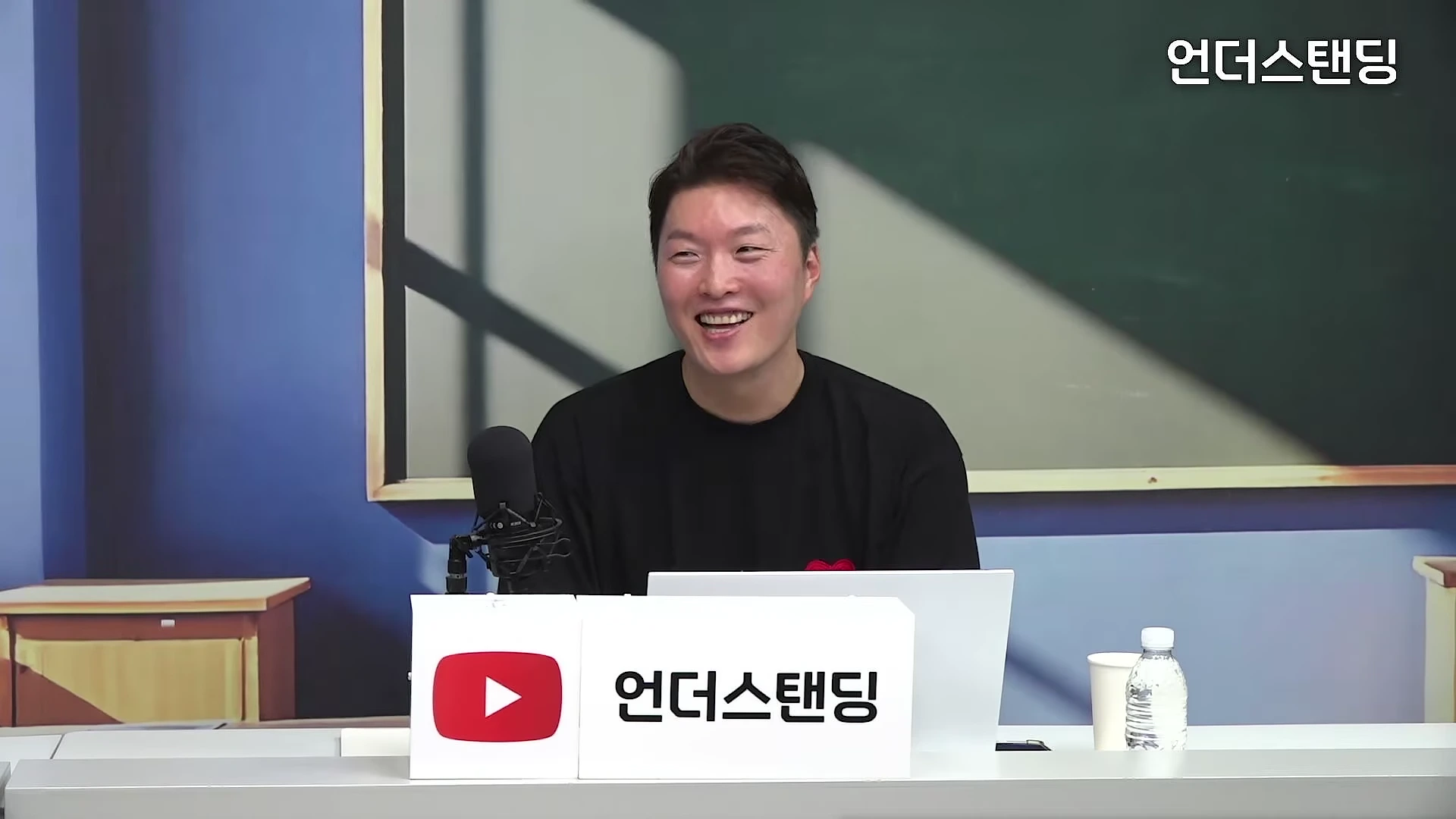 스튜디오에서 검은색 티셔츠를 입은 남성이 마이크 앞에 앉아 환하게 웃고 있는 모습