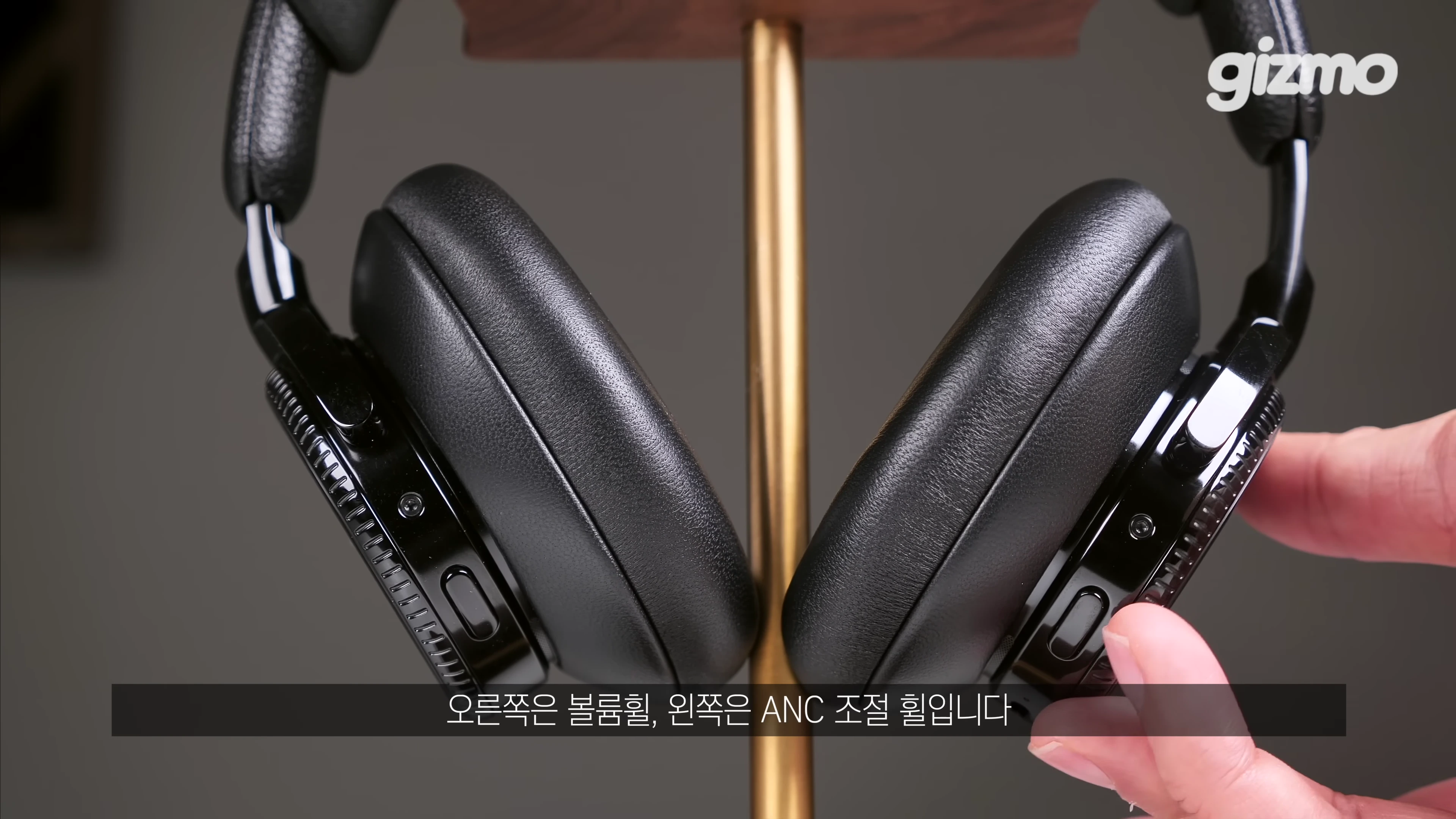 스탠드에 거치된 올블랙 헤드폰의 이어컵 측면 조작부를 손가락으로 가리키는 모습