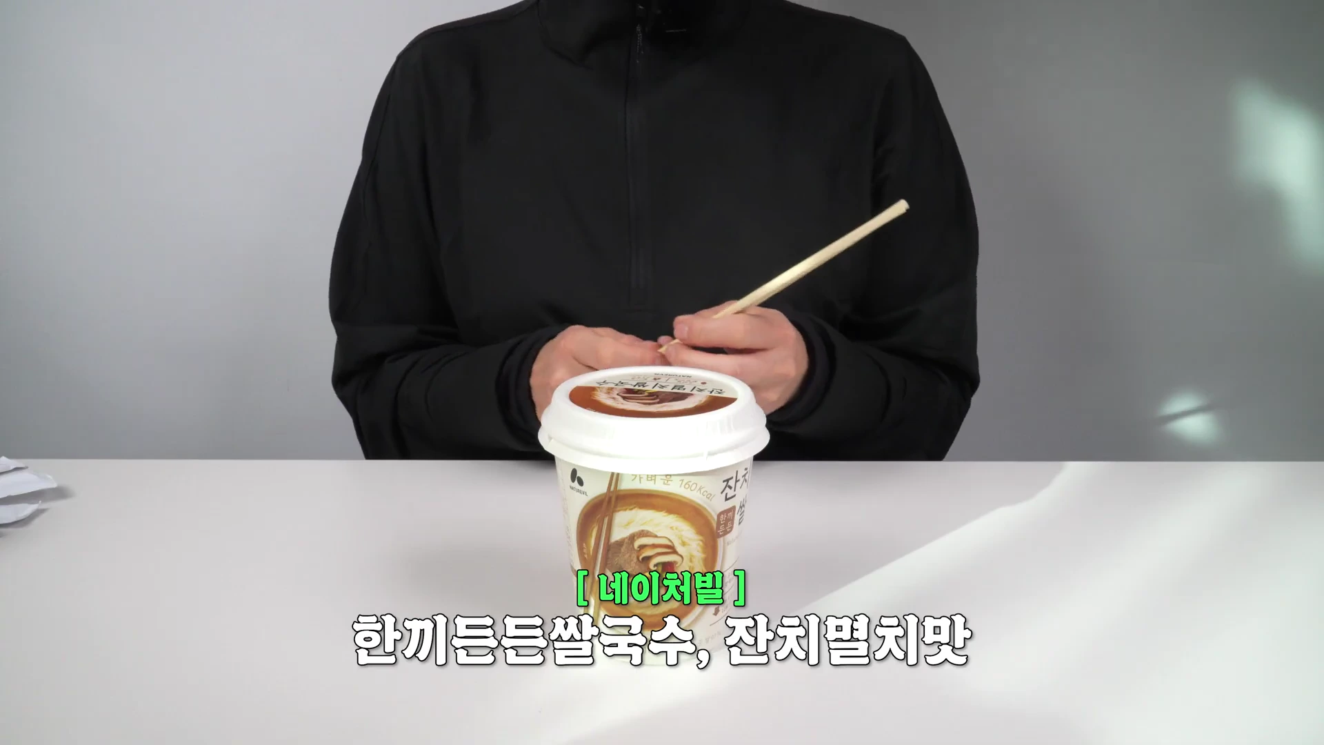 검은색 옷을 입은 사람이 테이블 위에 놓인 네이처빌 쌀국수 컵라면을 들고 있는 모습
