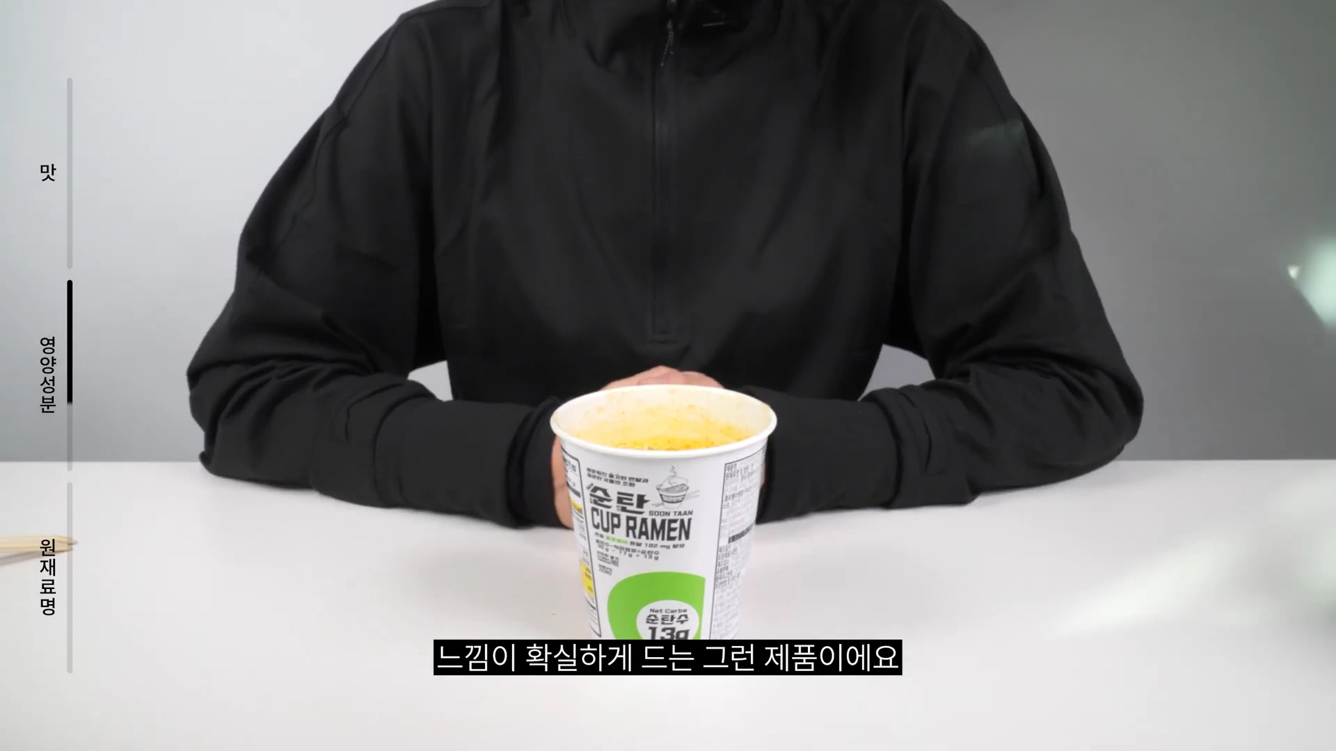 검은색 상의를 입은 사람이 테이블 위에 놓인 순탄 컵라면을 앞에 두고 앉아 있는 모습