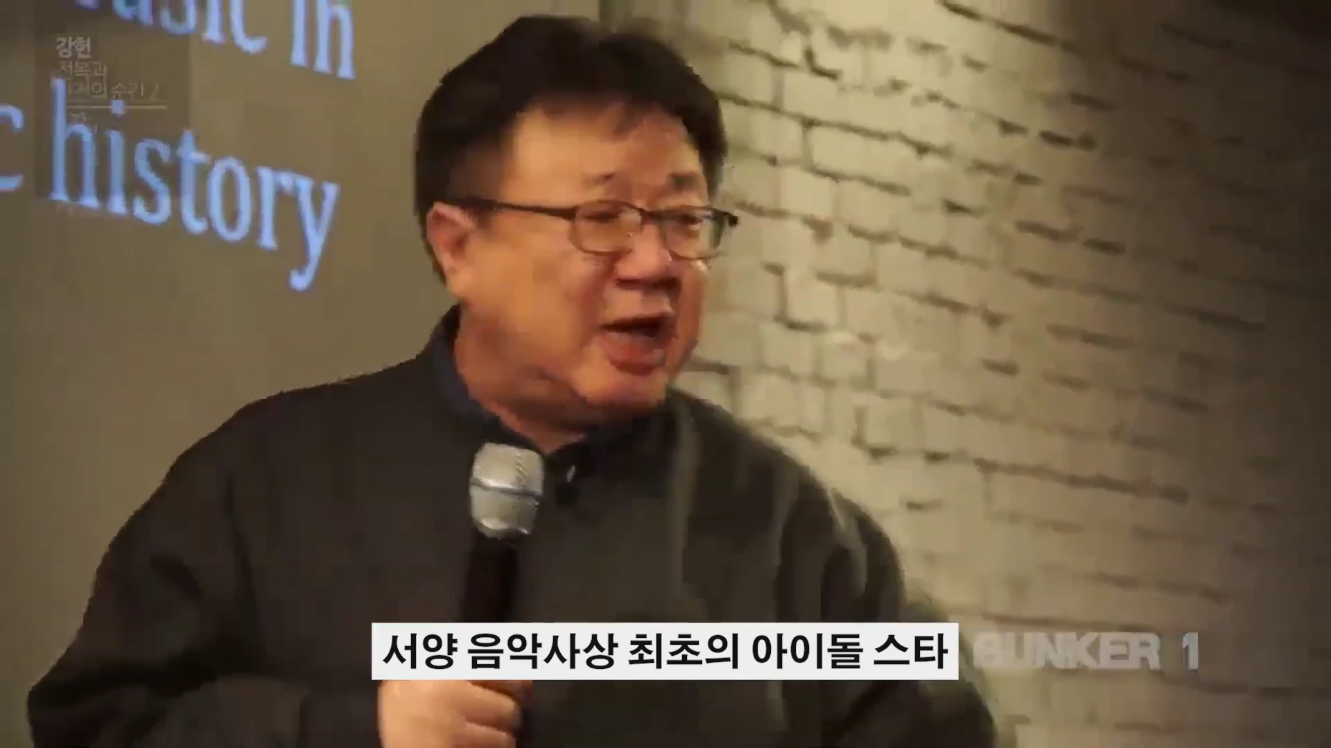 안경을 쓴 중년 남성이 마이크를 들고 강연하는 모습