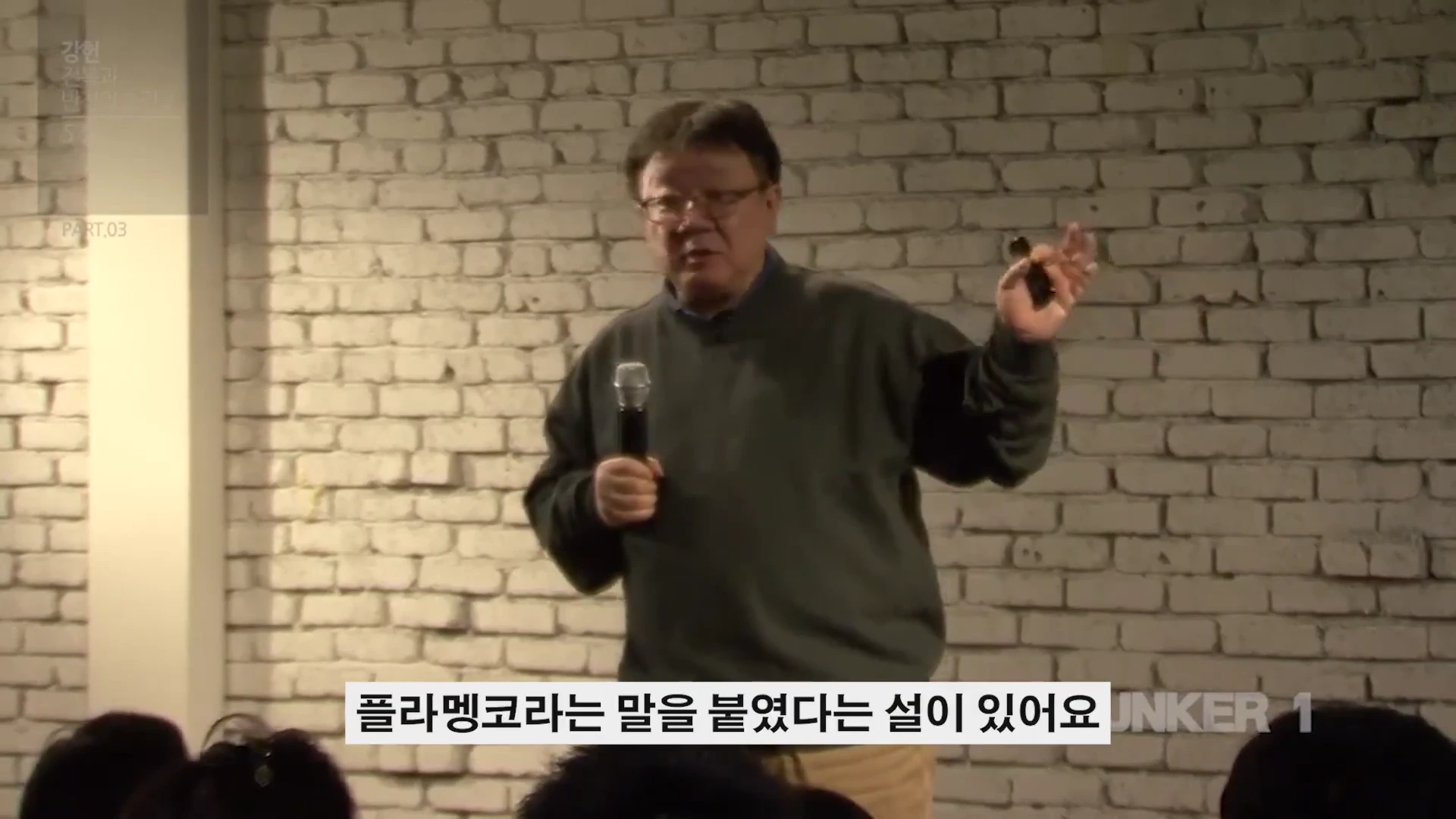 흰색 벽돌 배경 앞에서 마이크를 들고 강연하는 중년 남성