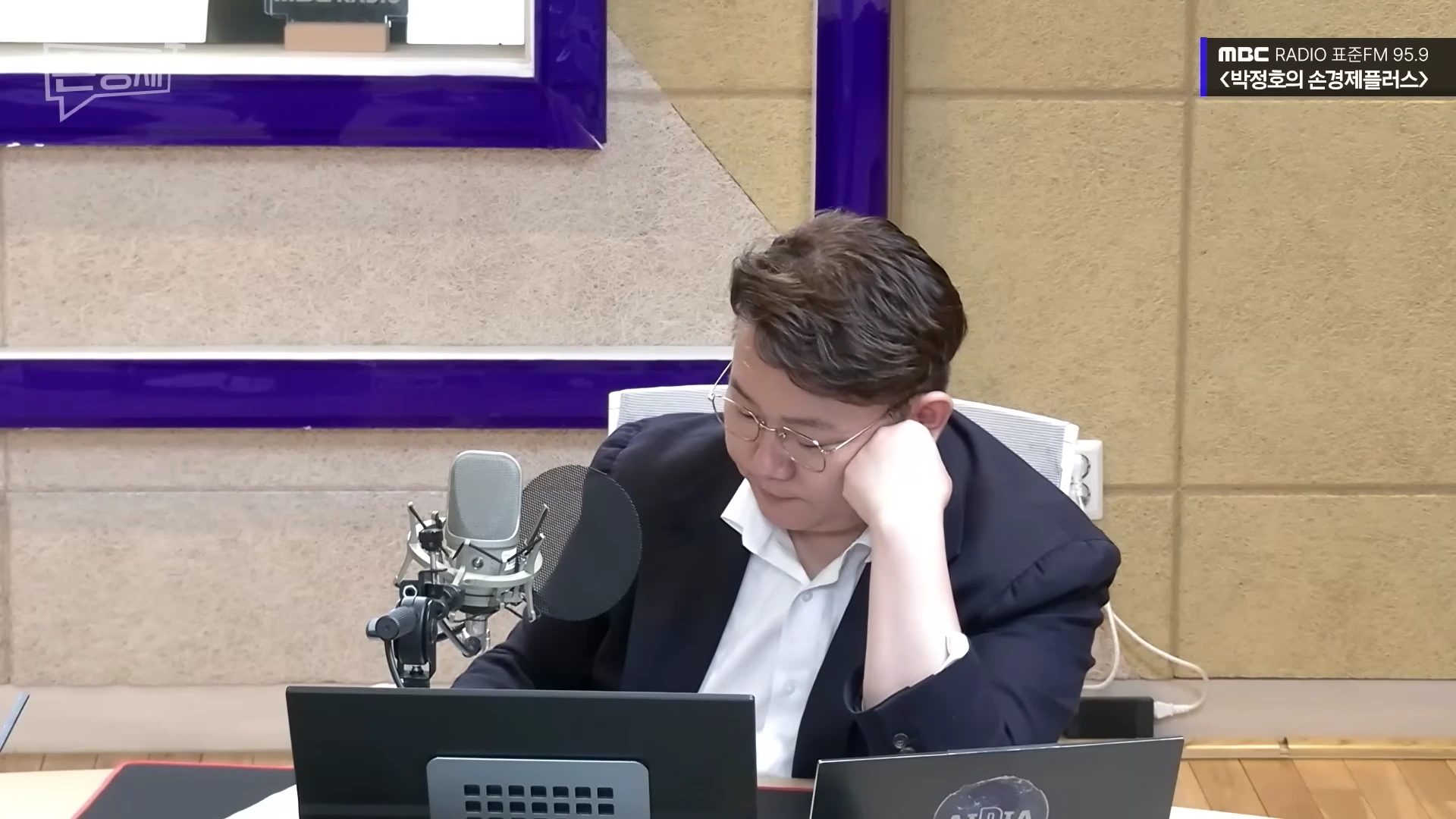 라디오 스튜디오에서 안경을 쓴 남성이 마이크 앞에 앉아 노트북 화면을 내려다보고 있다.