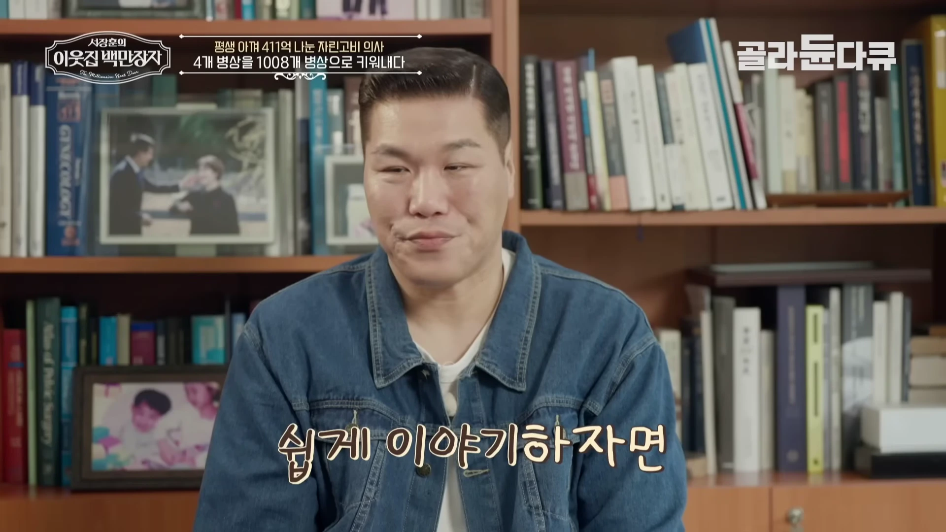 책장이 배경인 실내에서 데님 재킷을 입은 남성이 정면을 바라보며 이야기하는 모습