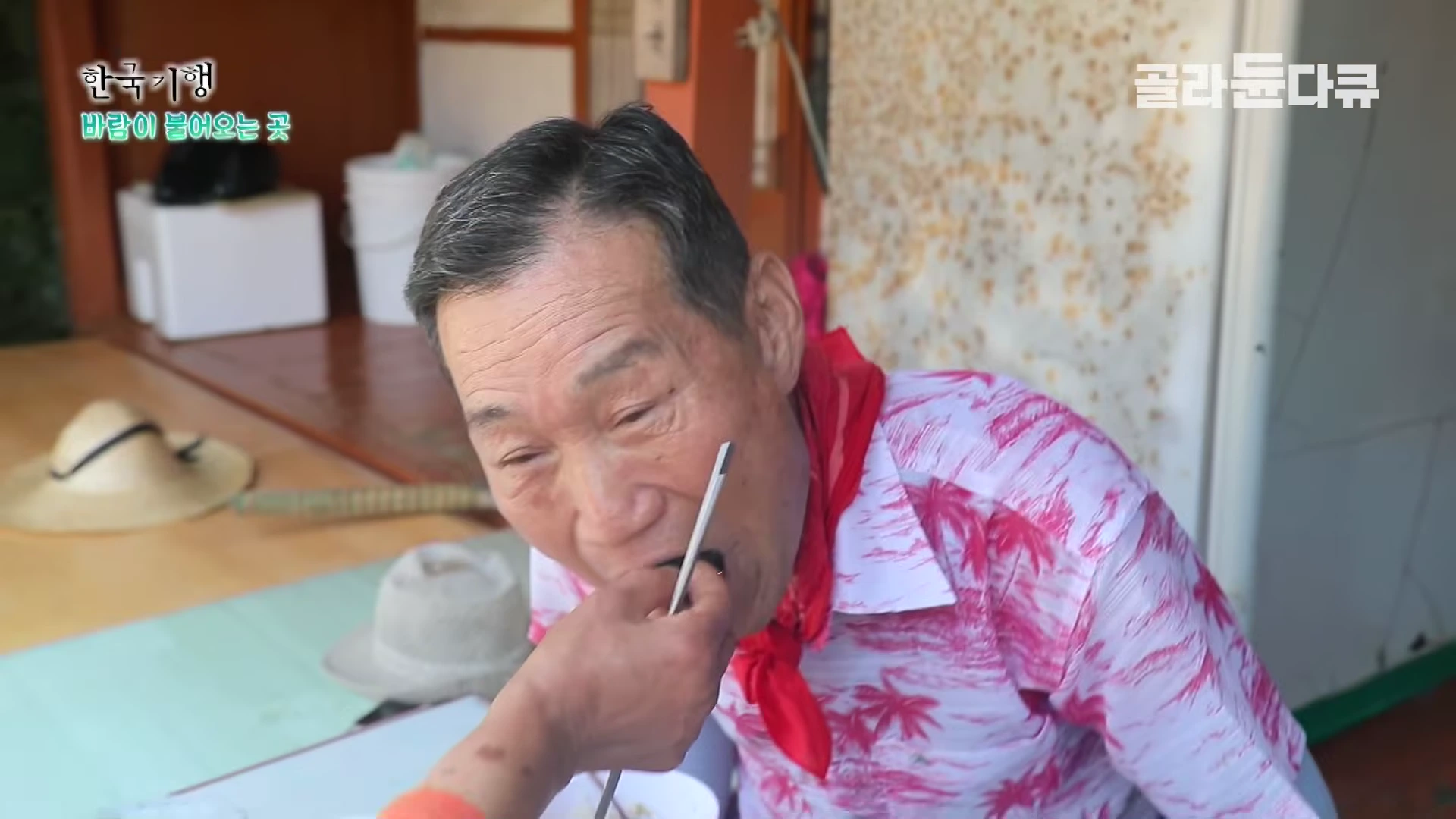 분홍색 무늬 셔츠를 입은 중년 남성이 누군가 건네주는 음식을 입으로 받아먹고 있다.