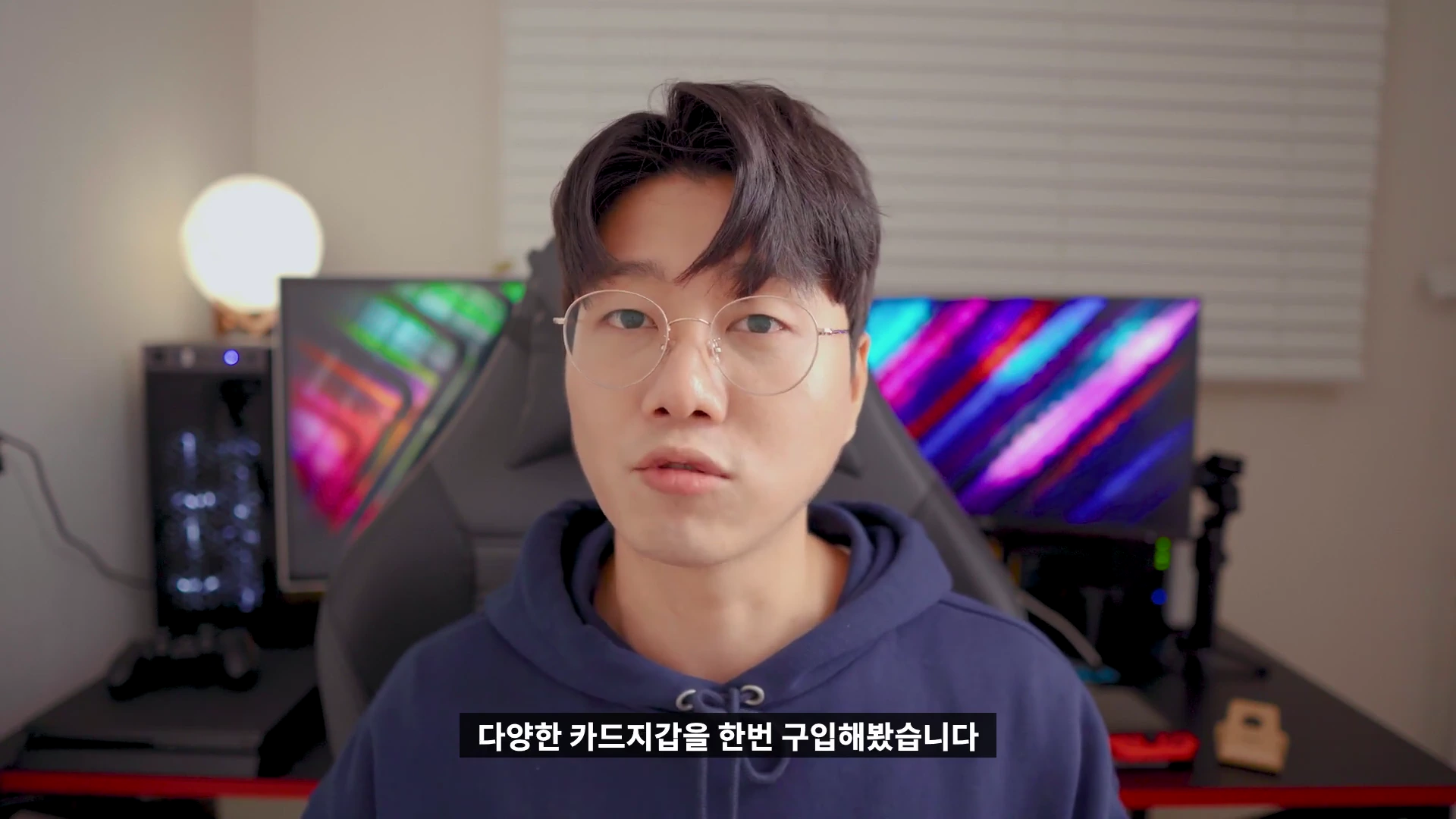 안경을 쓴 남성이 방 안에서 카메라를 정면으로 응시하며 이야기하고 있는 모습