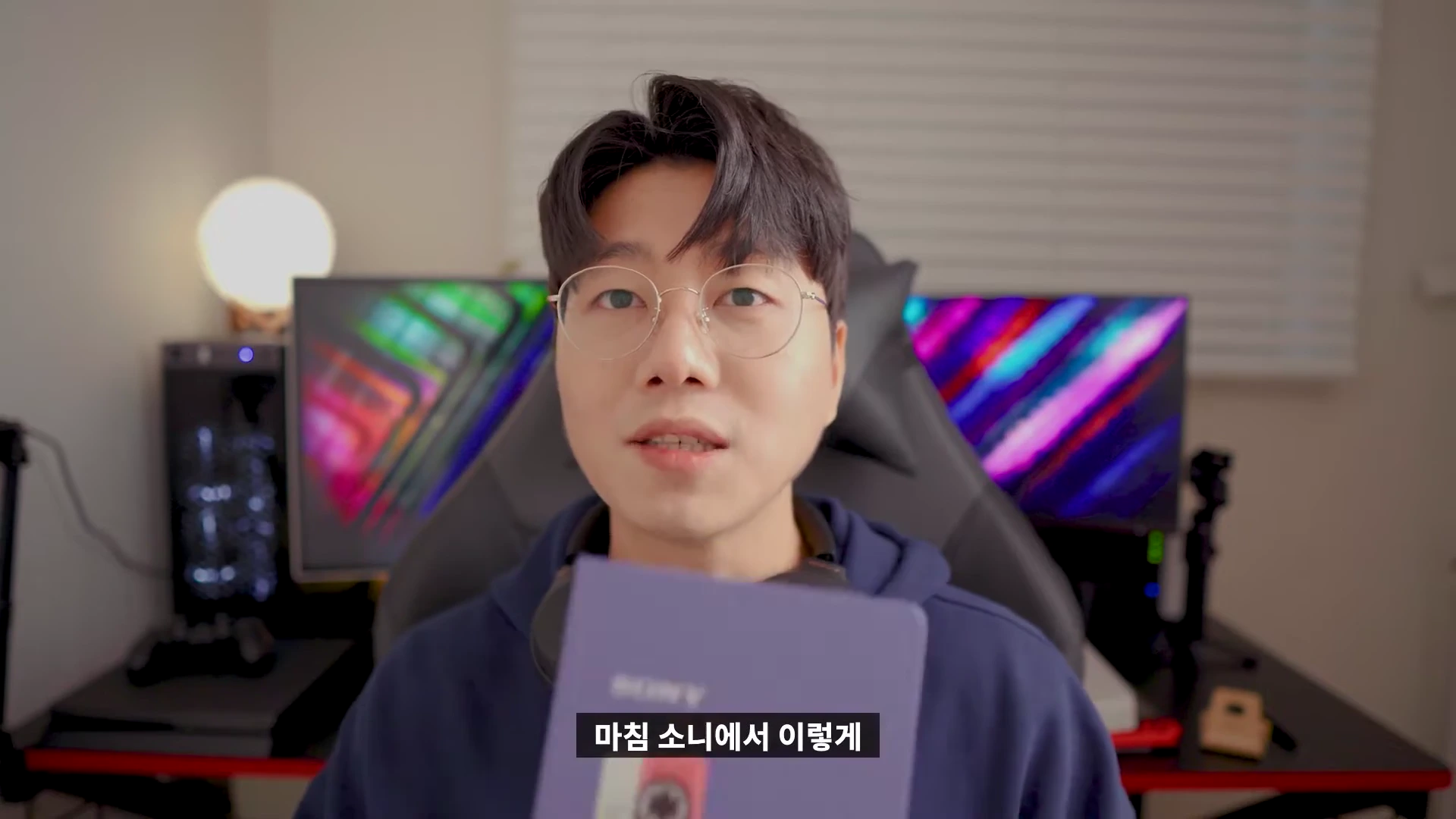 안경을 쓴 남성이 카메라를 보며 이야기하고 있고, 손에는 보라색 다이어리를 들고 있다.