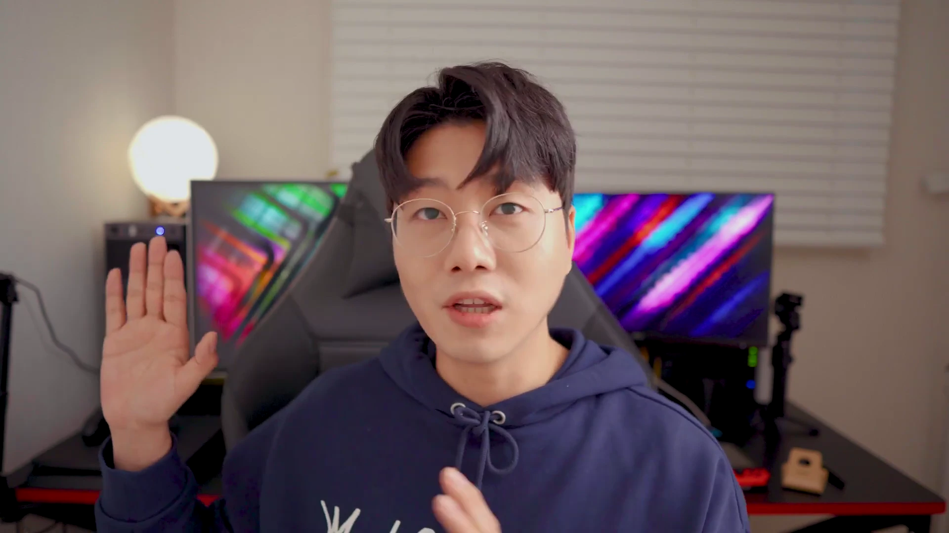 안경을 쓴 남성이 책상 앞에 앉아 손을 들어 인사하며 유튜브 영상을 촬영하는 모습