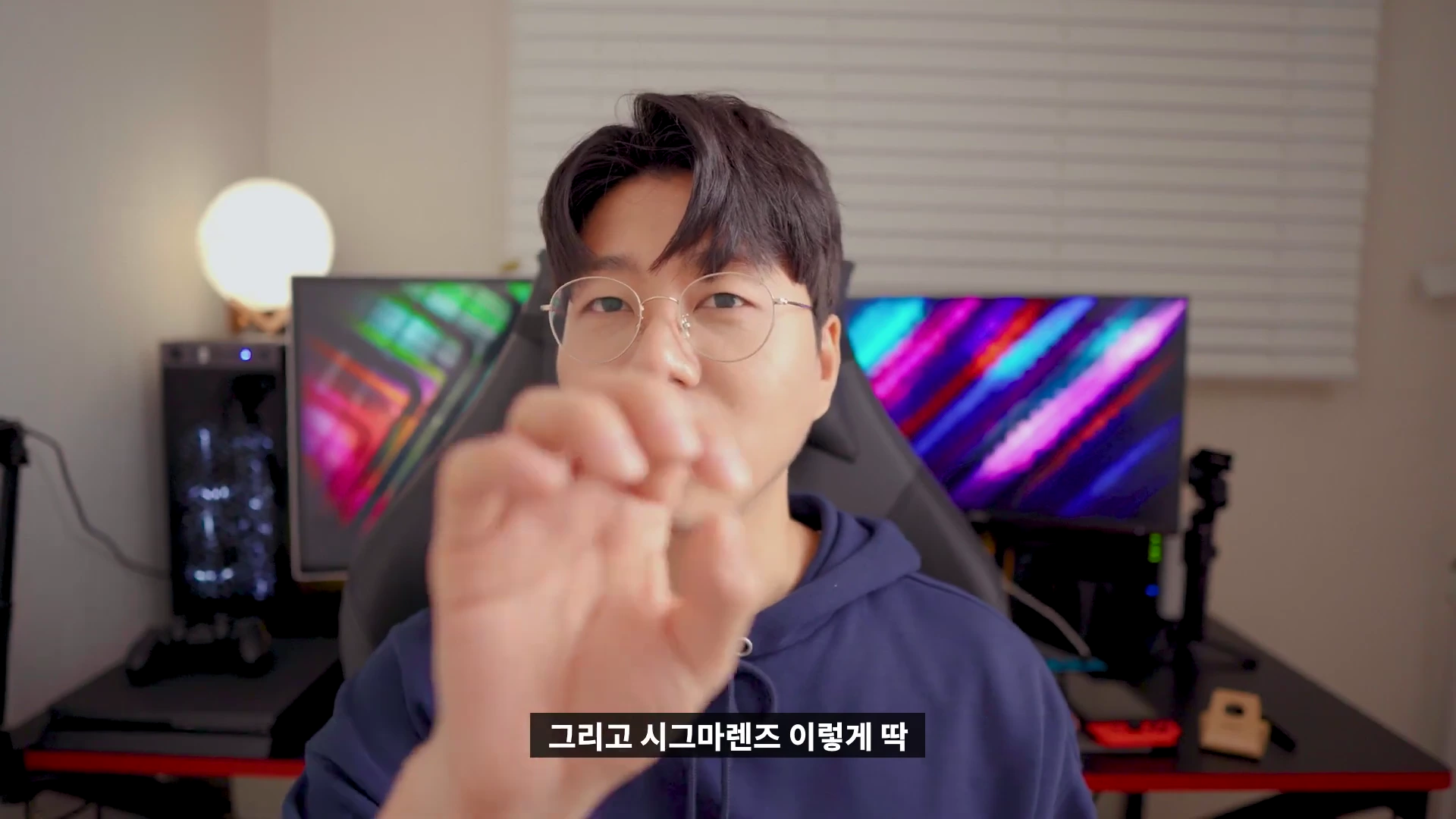 안경을 쓴 남성이 책상 앞에 앉아 손을 뻗어 카메라를 향해 제스처를 취하고 있는 모습