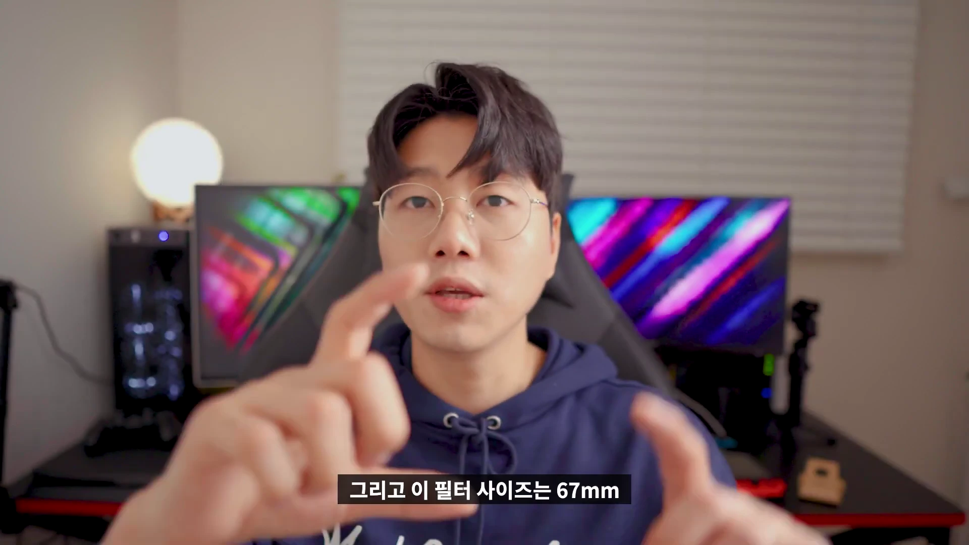 안경을 쓴 남성이 실내에서 카메라를 보며 손짓으로 설명하는 모습