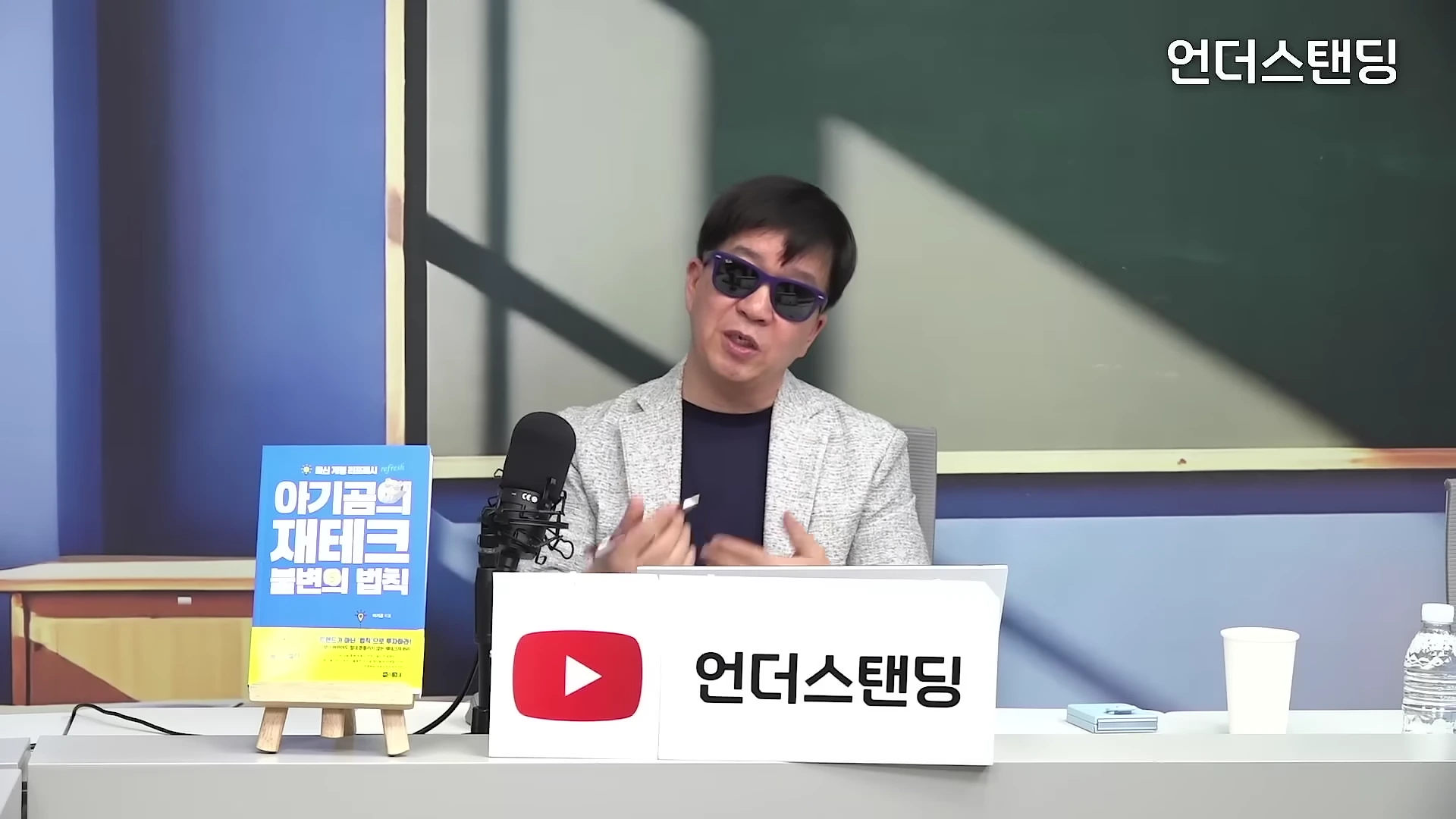 선글라스를 쓴 남성이 스튜디오 책상에 앉아 마이크를 앞에 두고 이야기를 나누고 있다.