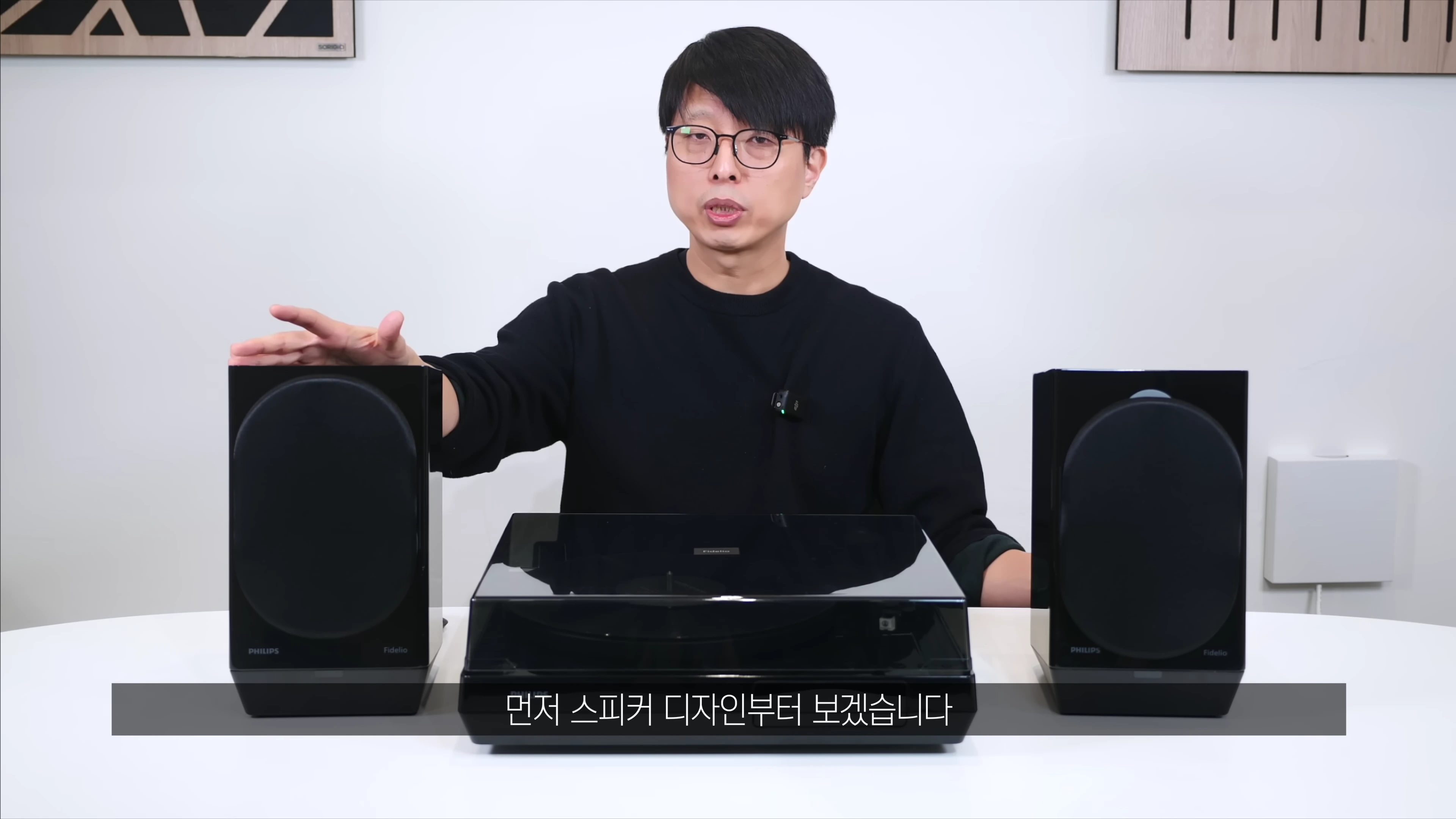 안경을 쓴 남성이 테이블 위에 놓인 턴테이블과 양옆의 스피커 세트를 소개하고 있다.