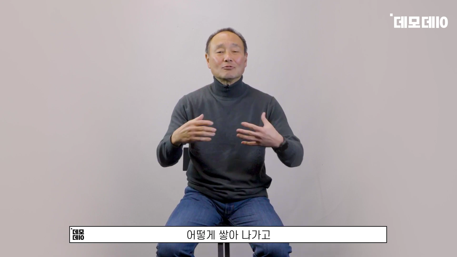 회색 터틀넥을 입은 중년 남성이 의자에 앉아 손짓하며 인터뷰하는 모습