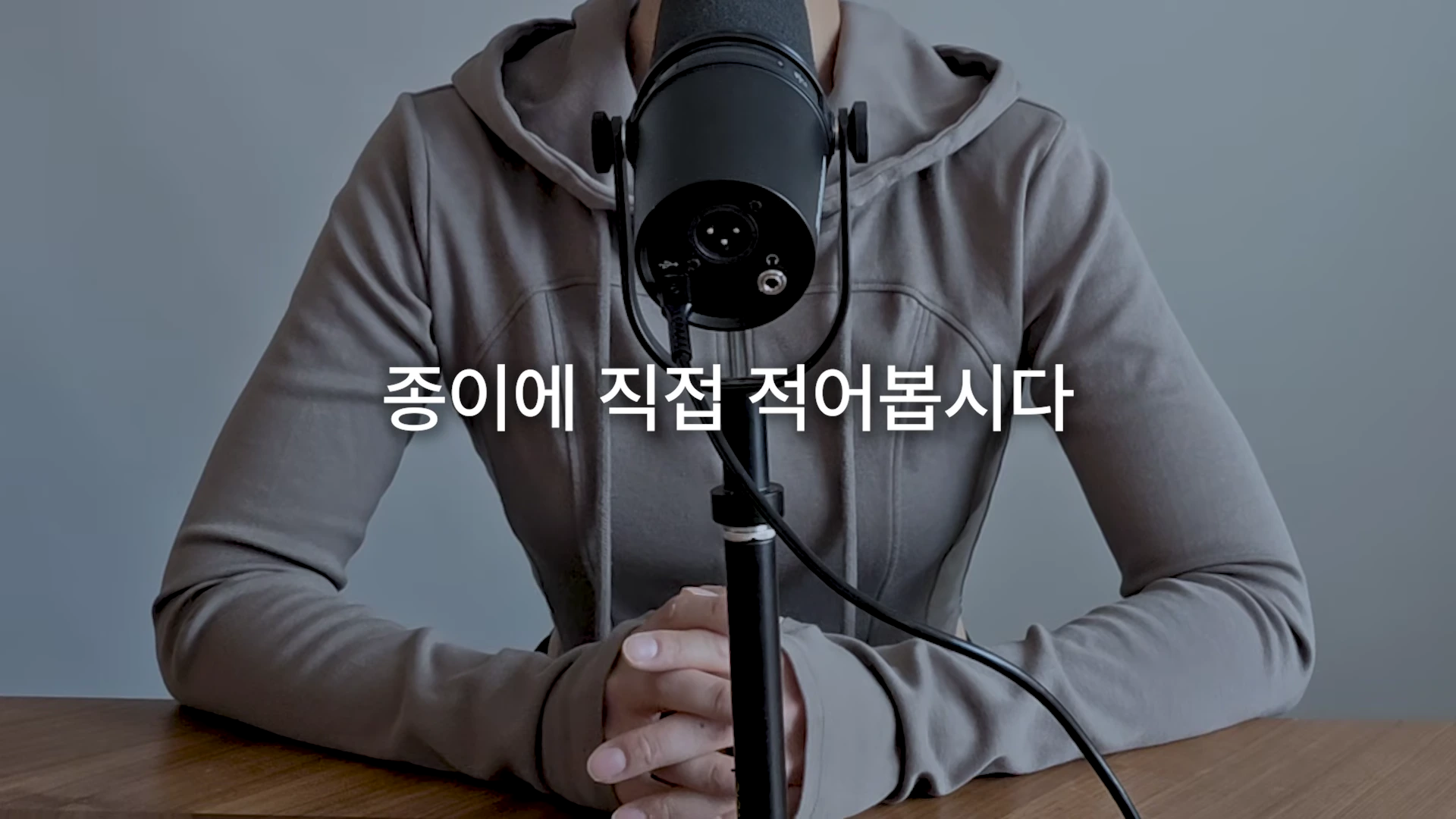 후드티를 입은 사람이 마이크 앞에 앉아 있고 화면 중앙에 '종이에 직접 적어봅시다'라는 흰색 자막이 떠 있다.
