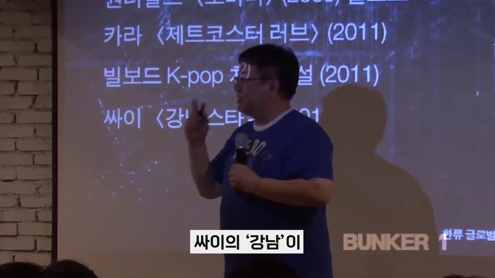 강연자가 마이크를 들고 빌보드 K-pop 차트 관련 내용이 적힌 스크린 앞에서 발표하고 있다.