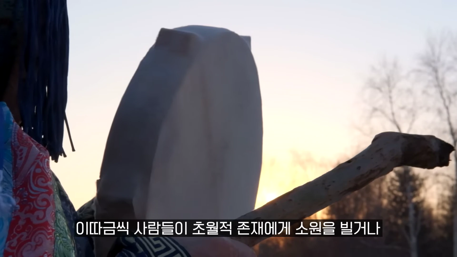 전통 의상을 입은 사람이 북과 북채를 들고 노을을 배경으로 서 있는 모습