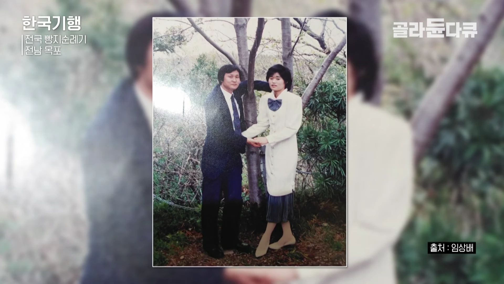 나무 옆에 나란히 서서 손을 맞잡고 있는 젊은 시절 부부의 옛날 사진