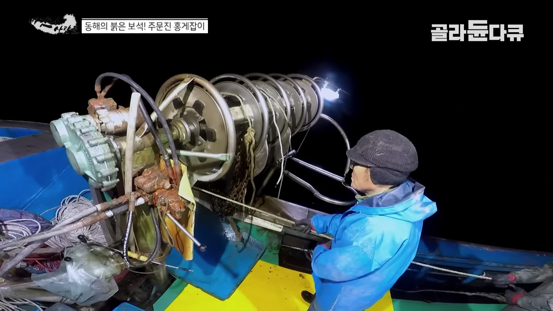 어두운 밤바다 위 어선에서 작업자가 기계 장치를 이용해 긴 밧줄과 그물을 끌어올리고 있다.