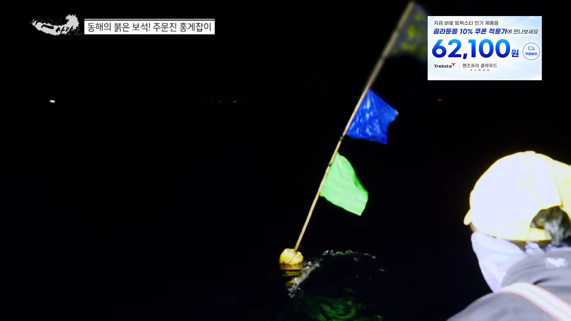 어두운 밤바다 위로 파란색과 초록색 깃발이 달린 부표가 떠 있고, 노란 모자를 쓴 선원이 이를 바라보고 있다.