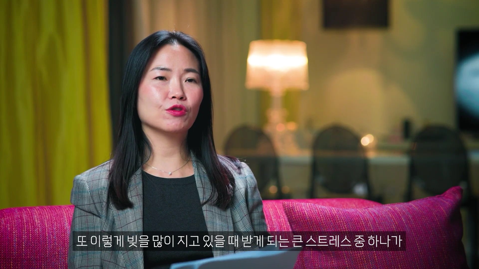체크무늬 재킷을 입은 여성이 분홍색 소파에 앉아 인터뷰하는 모습