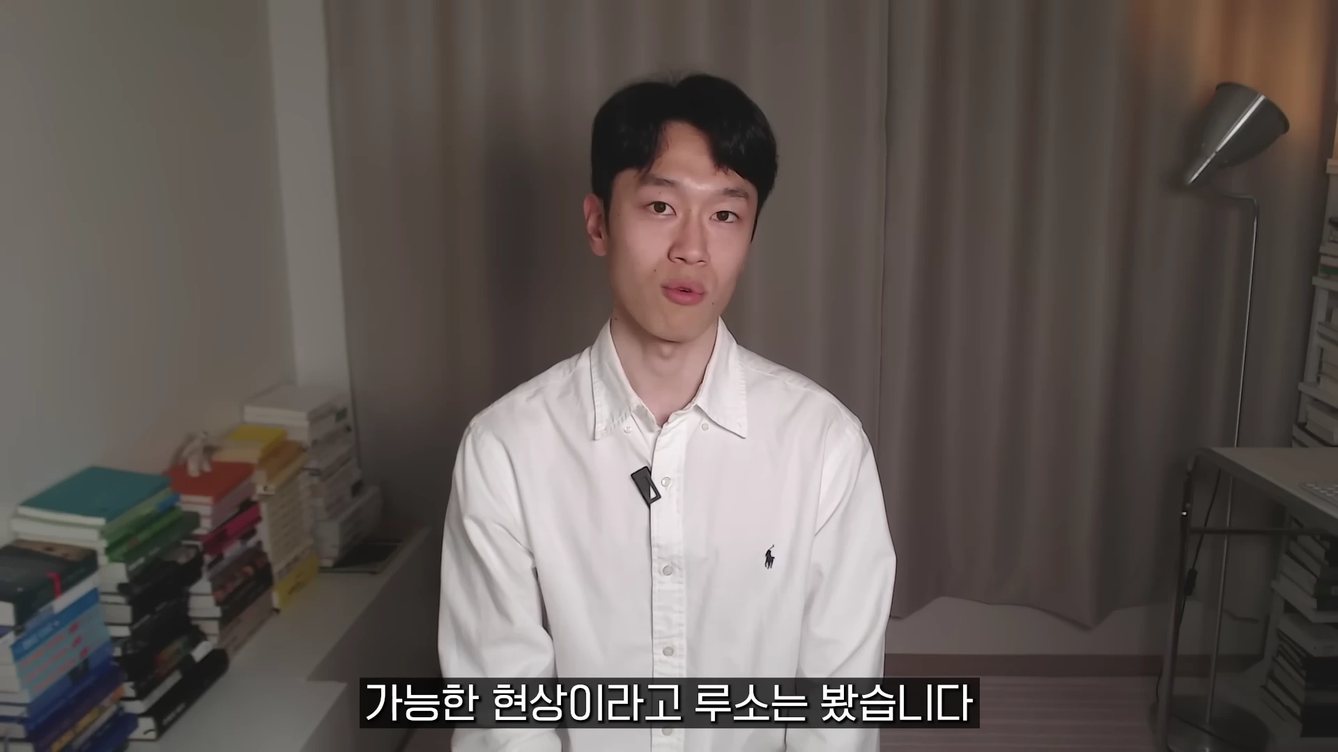흰 셔츠를 입은 남성이 실내에서 카메라를 응시하며 이야기하고 있다.