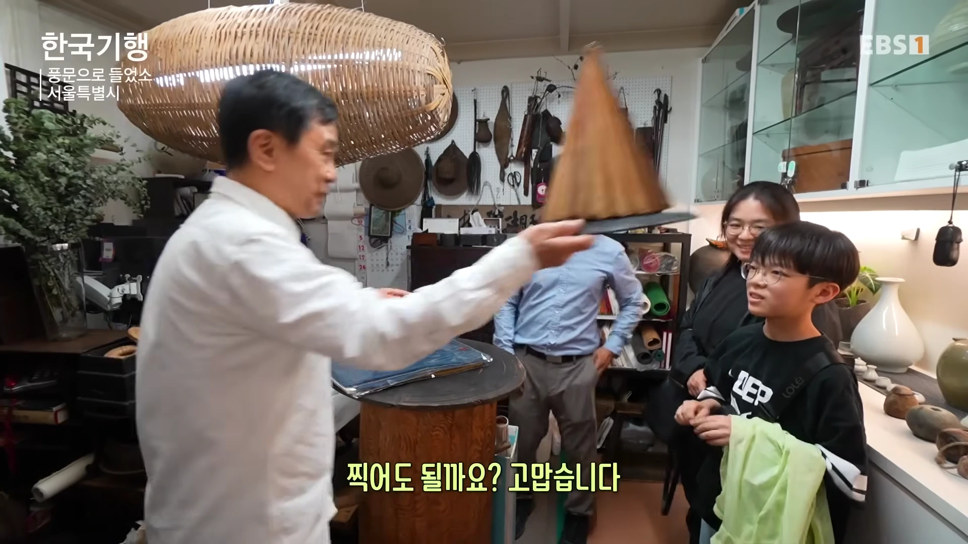 고미술품 가게에서 중년 남성이 아이에게 옛 모자인 갈모를 보여주고 있고, 그 뒤로 가족들이 호기심 어린 표정으로 바라보고 있다.